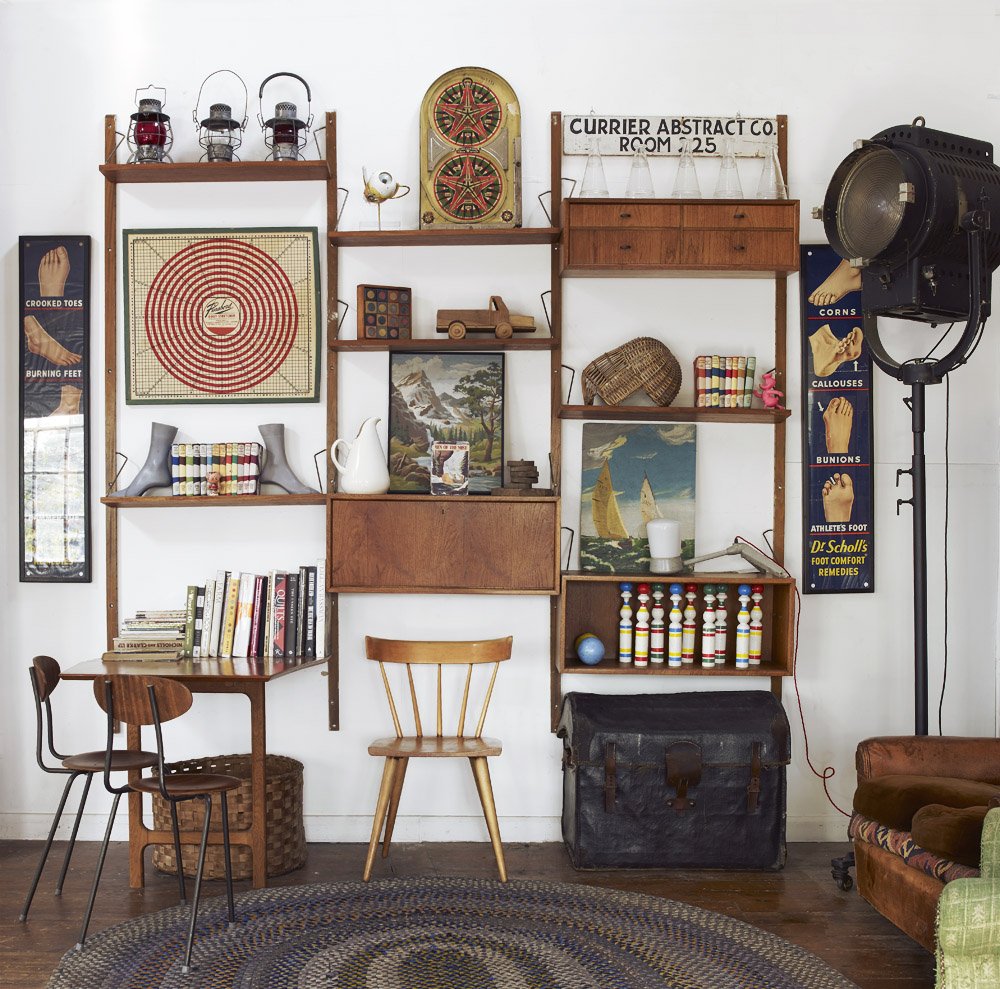 Decorative wall with wooden shelves holding vintage and eclectic items including framed pictures, a model car, small dolls, books, and a sphere, adjacent to vintage posters about foot health, and a large studio light on the right.
