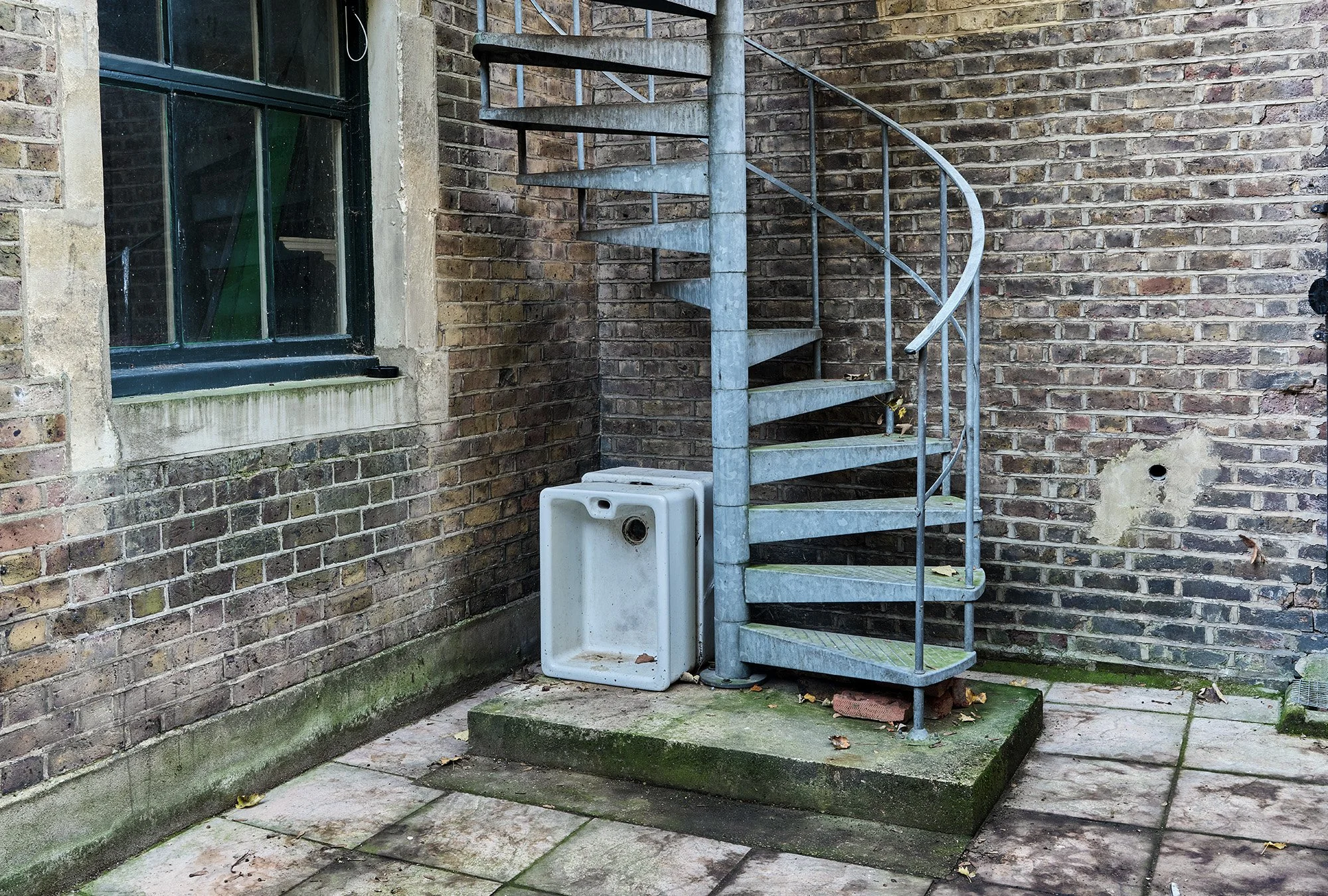 A metal spiral staircase beside a brick building wall with a window. There is a white sink or basin on the ground next to the staircase. The area has moss and dirt on the pavement.
