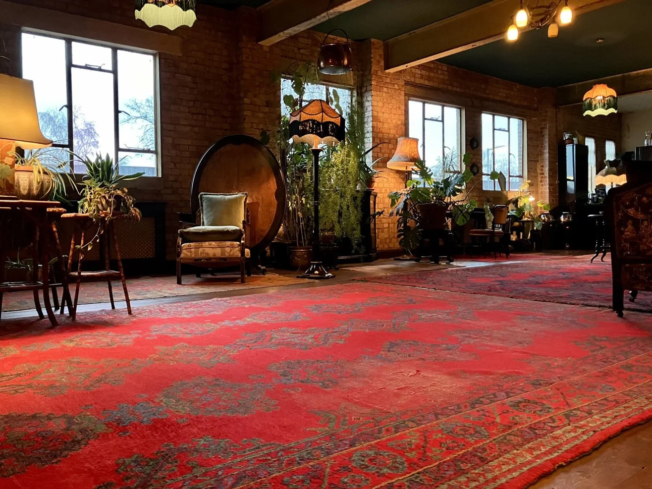 Cozy living room with brick walls, large windows, and vintage furniture. Persian rug, plants, and warm lighting create a welcoming atmosphere.
