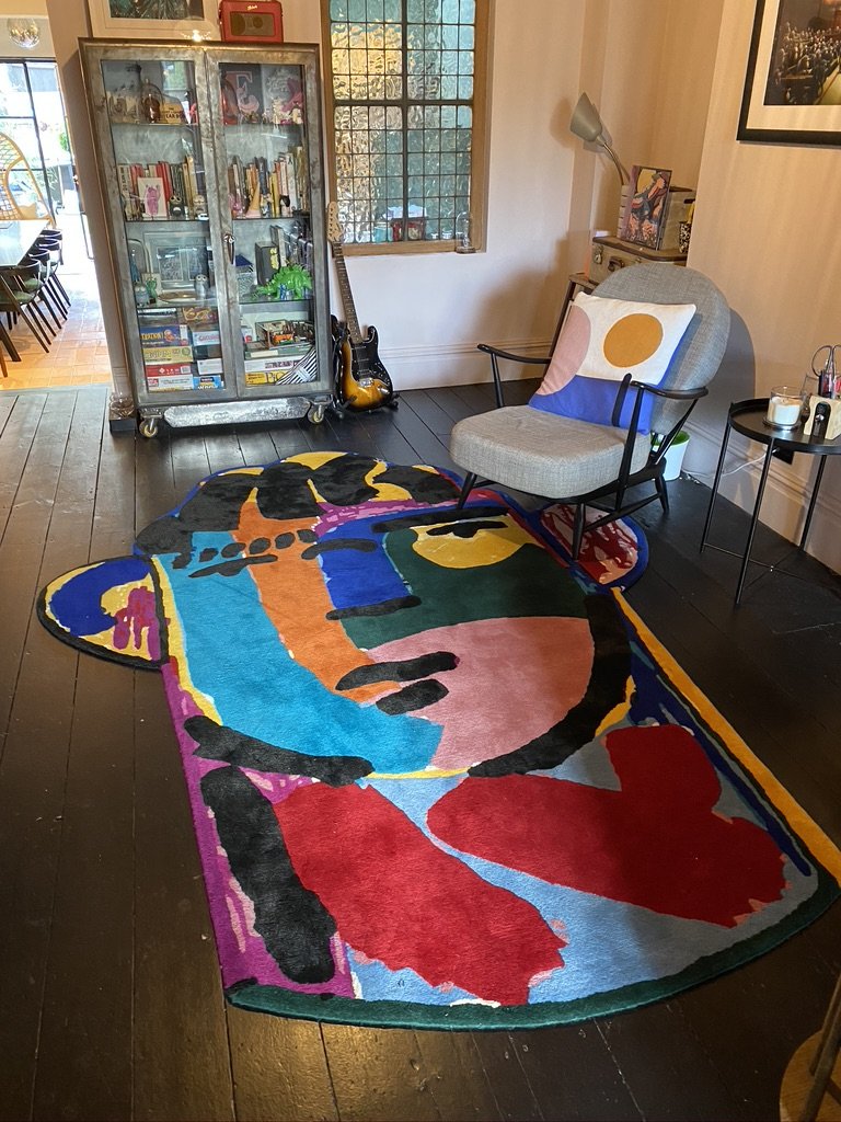 Colorful abstract rug on dark hardwood floor inside a cozy living room. A light gray armchair with a cushion sits beside a small black side table with candles, against a wall with a framed picture. A glass cabinet filled with books and decorative ite