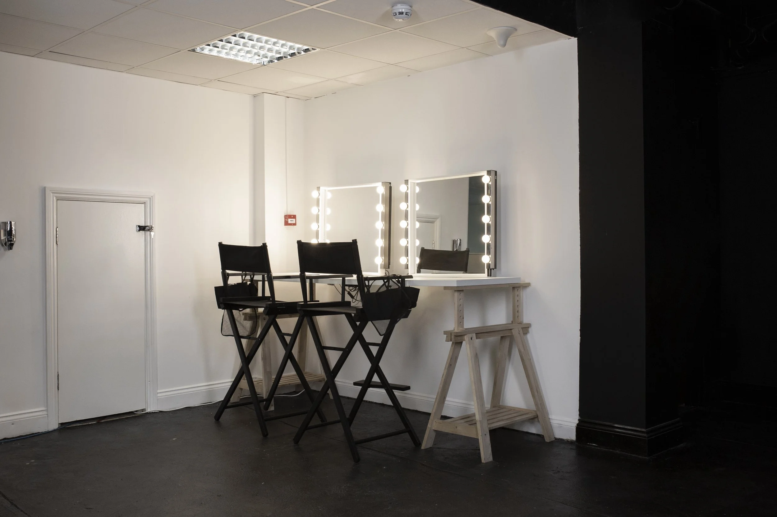 A makeup station with two illuminated mirrors, two black director's chairs, and a white table on a wooden stand, set against a white wall with a black section on the right. The ceiling has fluorescent lights and a smoke detector.