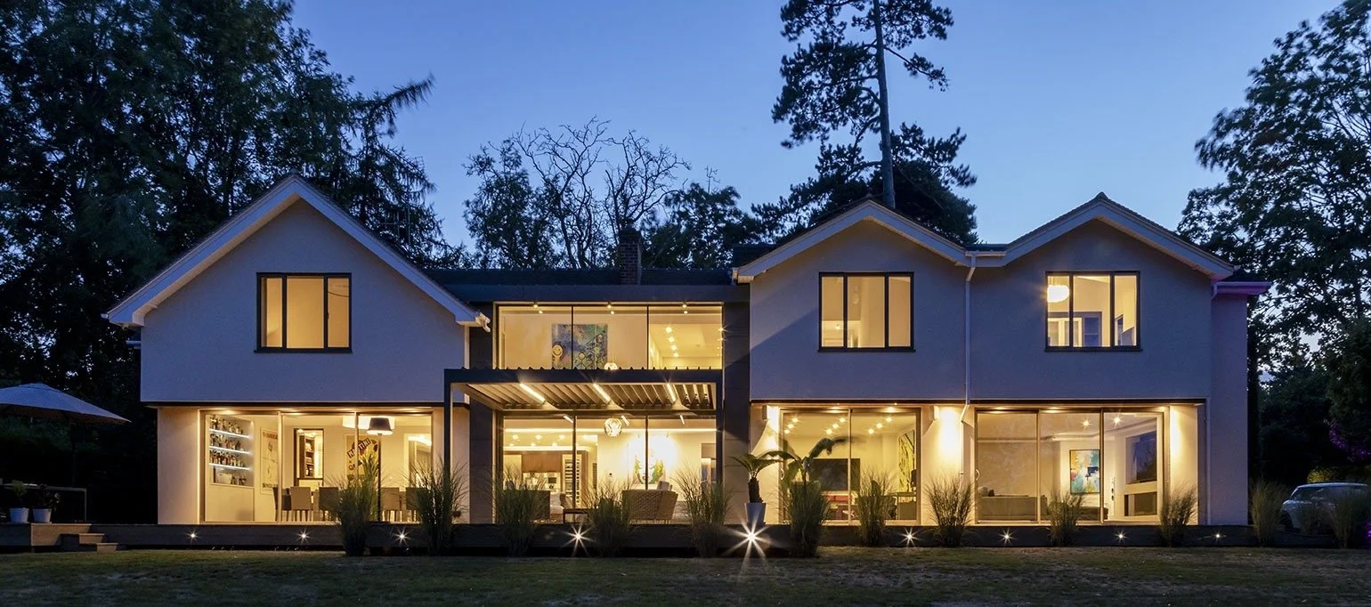 A two-story modern house at dusk with large illuminated glass windows and sliding doors, surrounded by trees and a landscaped yard.
