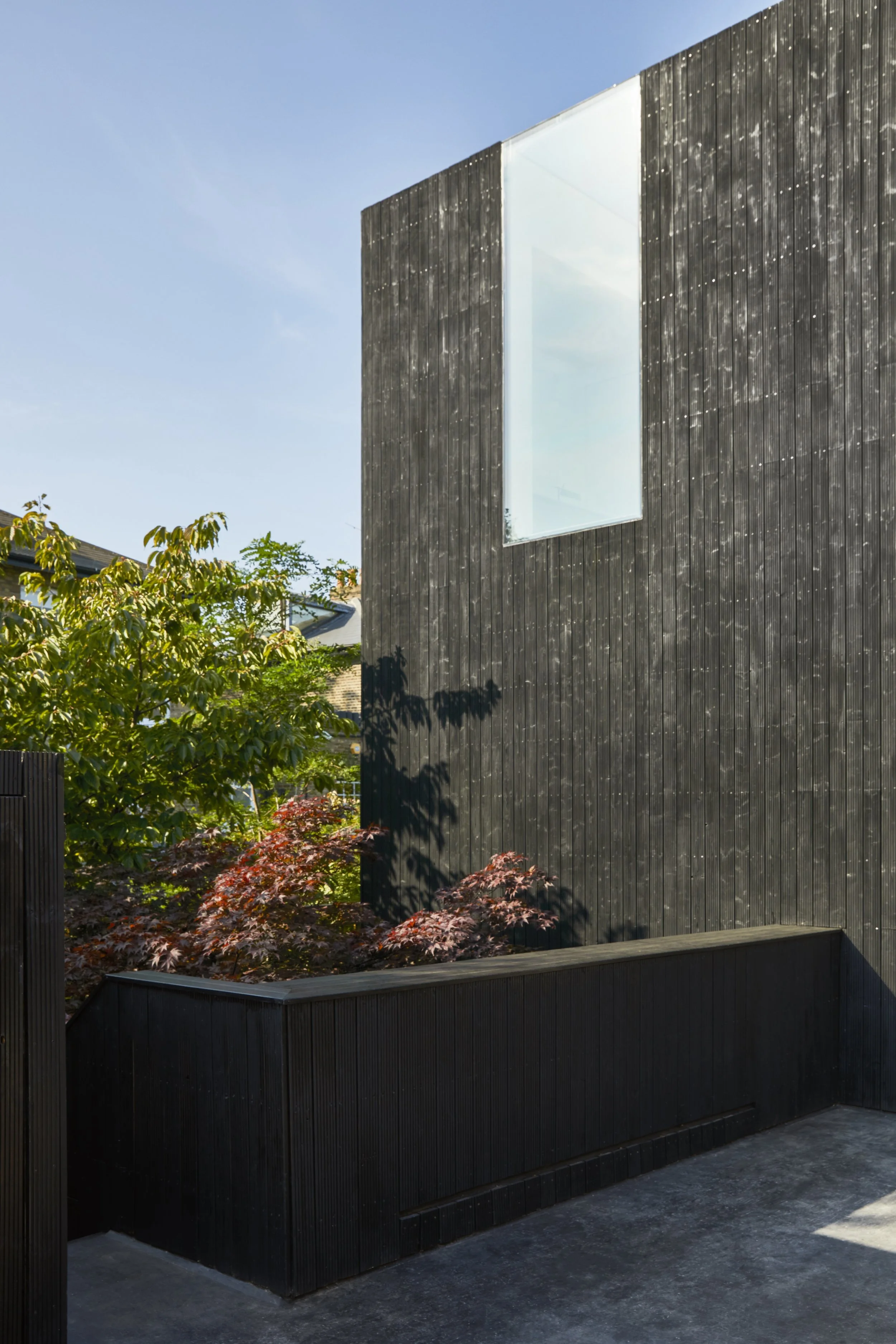 Modern black wooden building with a large rectangular window and lush green trees and shrubs nearby.