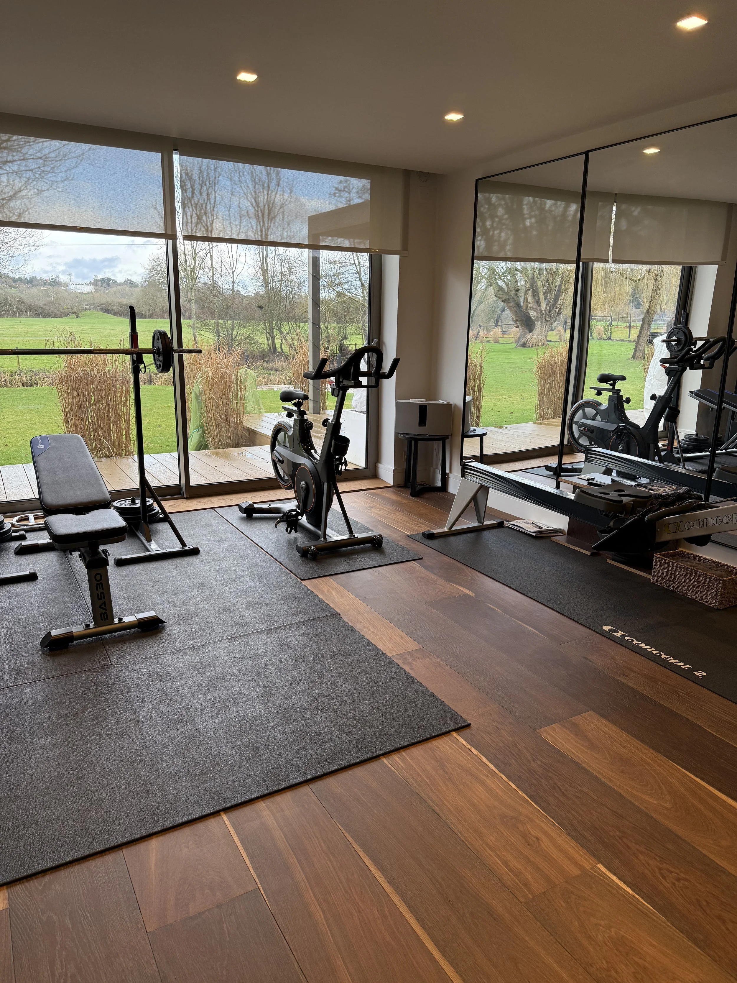Home gym with large windows overlooking a green outdoor landscape, featuring exercise equipment including a bench, stationary bike, and a rowing machine on a wooden floor with mats.