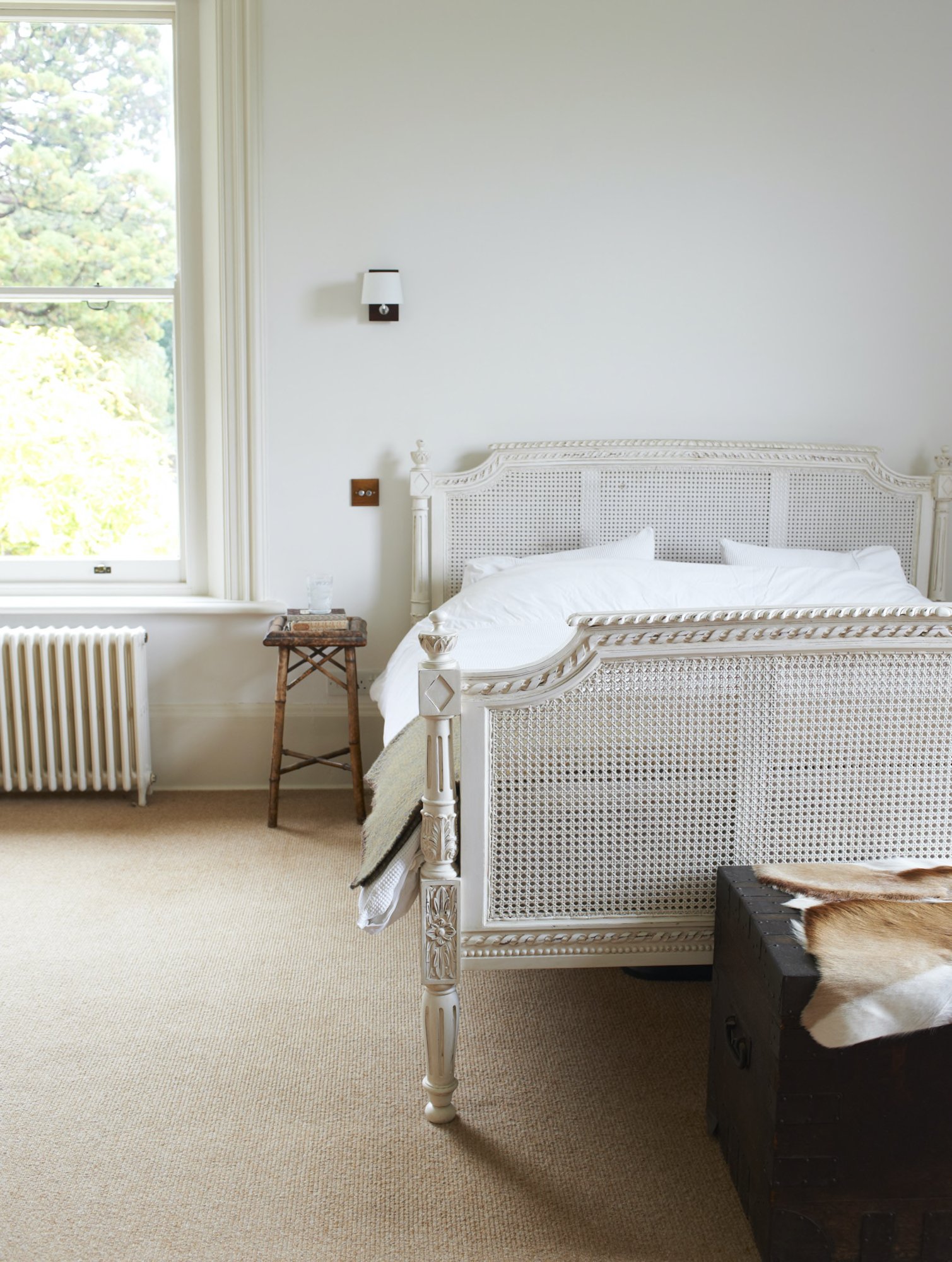 A bedroom with a window, a white carved bed with white bedding, a small wooden side table with a glass, a black chest with animal fur, and a beige carpet.