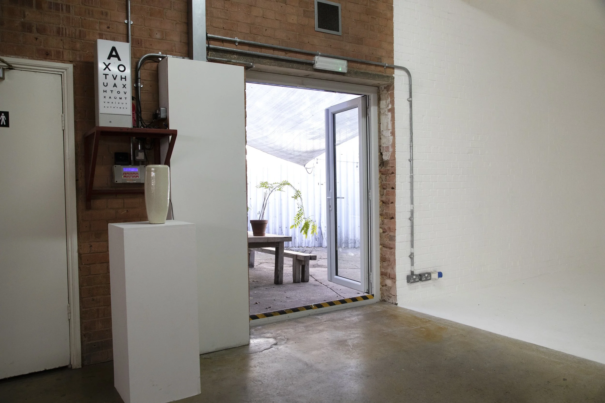 Interior of a gallery or store with white walls, an open door leading outside to a patio with plants, a pedestal with a white vase, an eye chart, and electrical wiring along the wall.