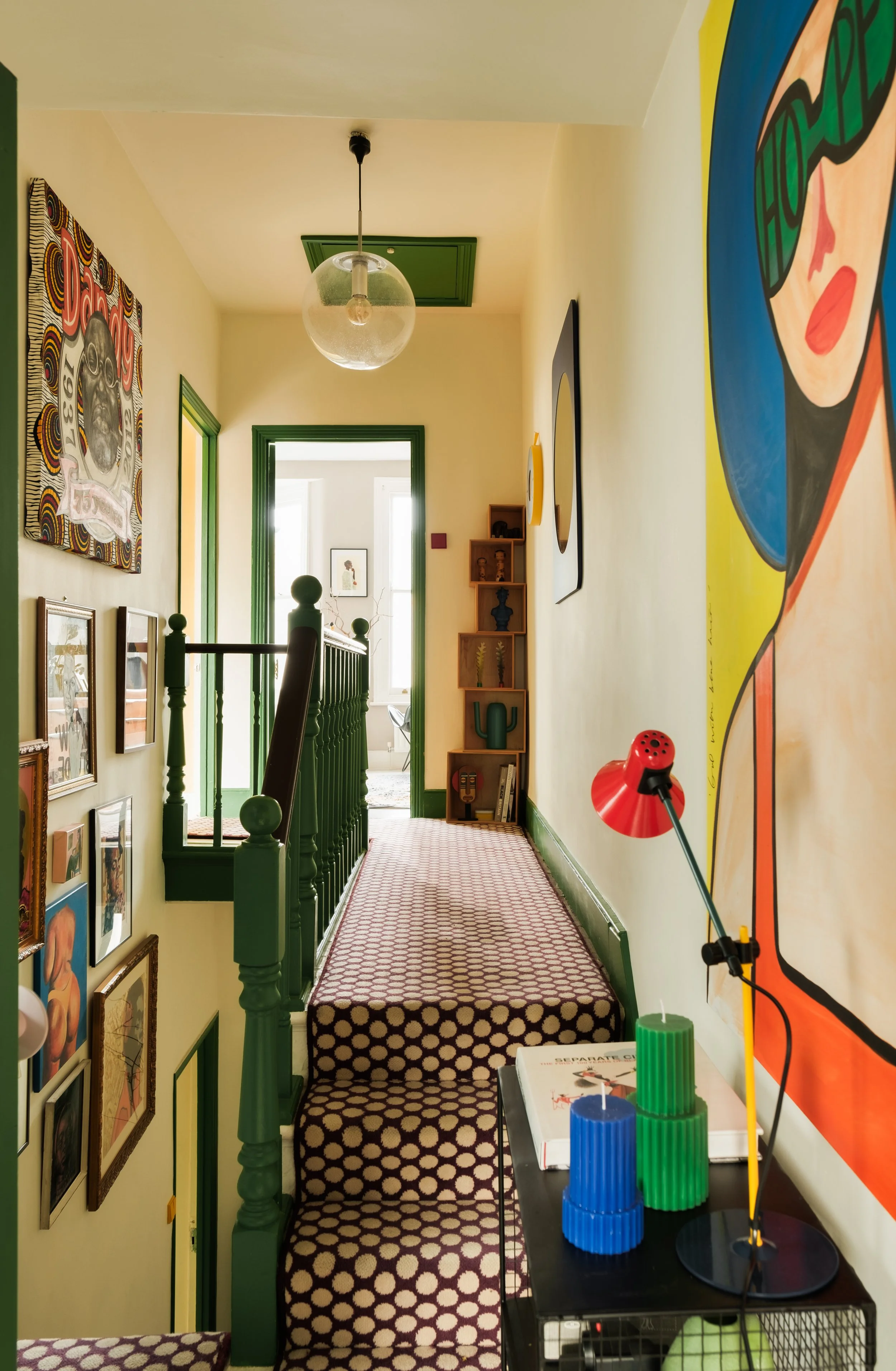 A hallway with yellow walls, green door and window trims, and patterned carpeted stairs. Decor includes framed art on the wall, a small wooden corner shelf with decorative vases and figurines, and a colorful large poster or painting. A black table ho