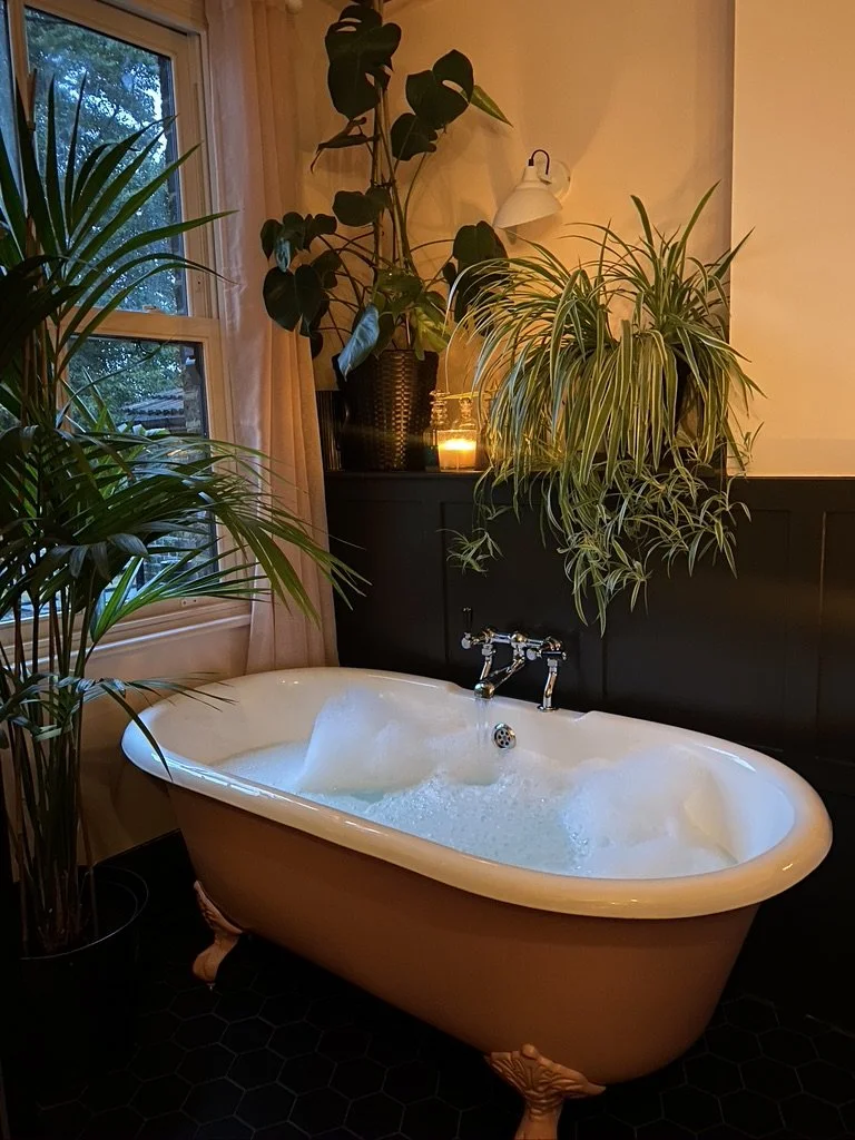 A vintage clawfoot bathtub filled with bubbly water in a cozy bathroom with plants, a candle, and a window.
