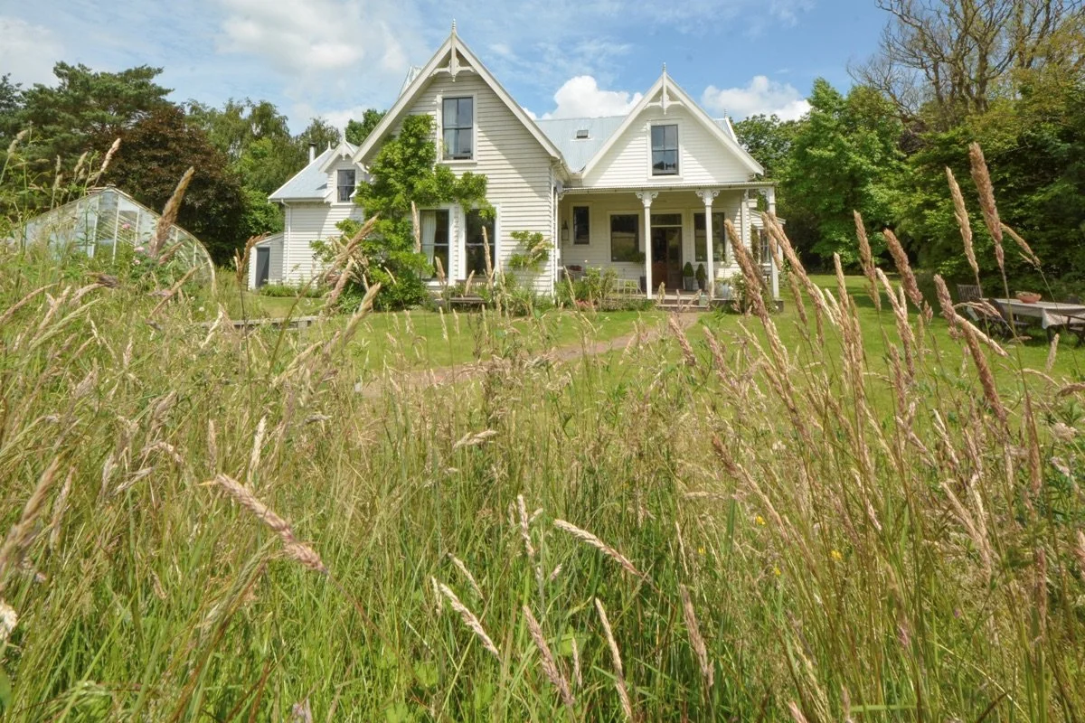 A white Victorian-style house with a porch, surrounded by trees and tall grass, with a greenhouse on the left and a picnic table on the right.