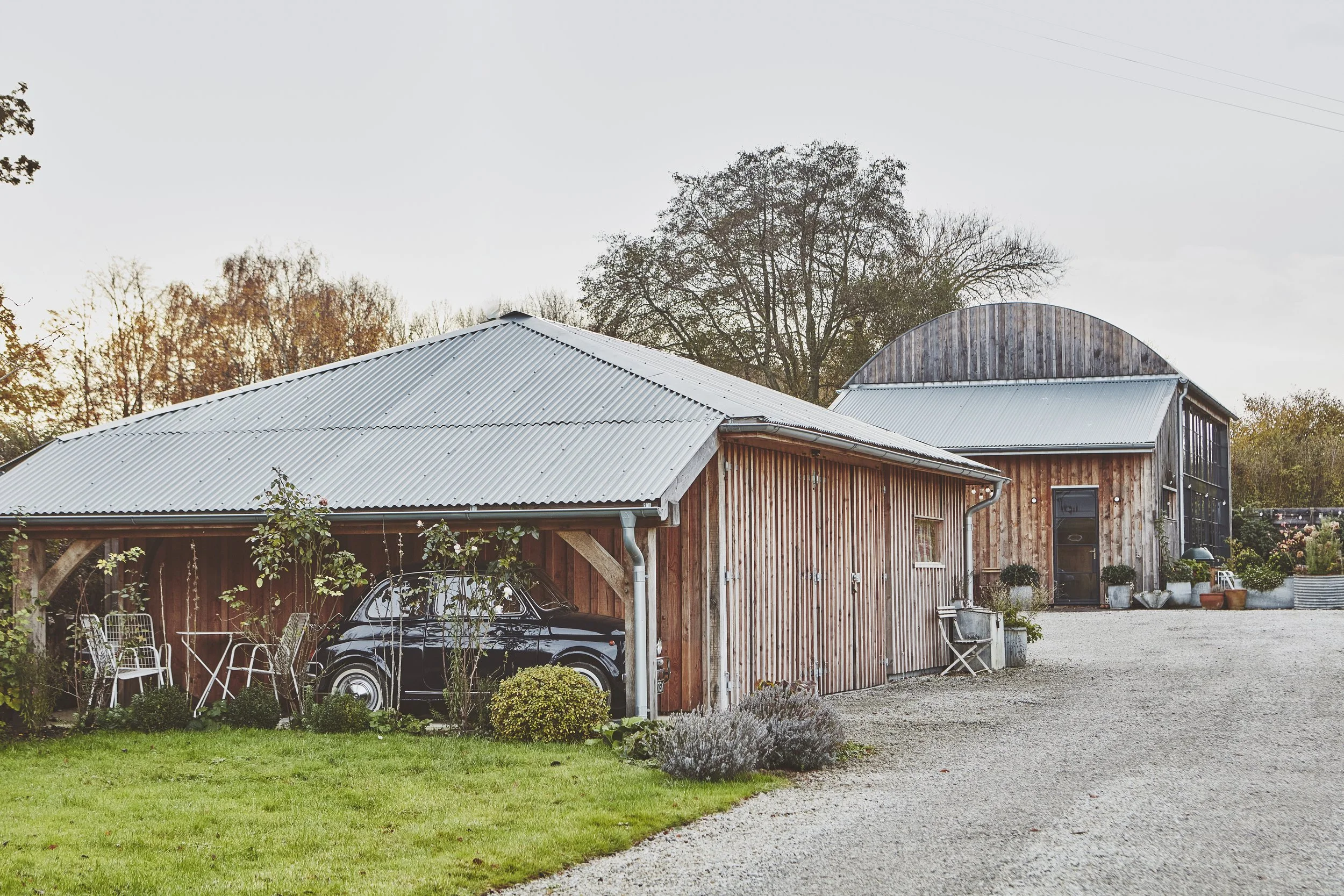 A rustic barn with a metal roof, wooden siding, and large windows, surrounded by a gravel driveway, green grass, and potted plants, with trees in the background.