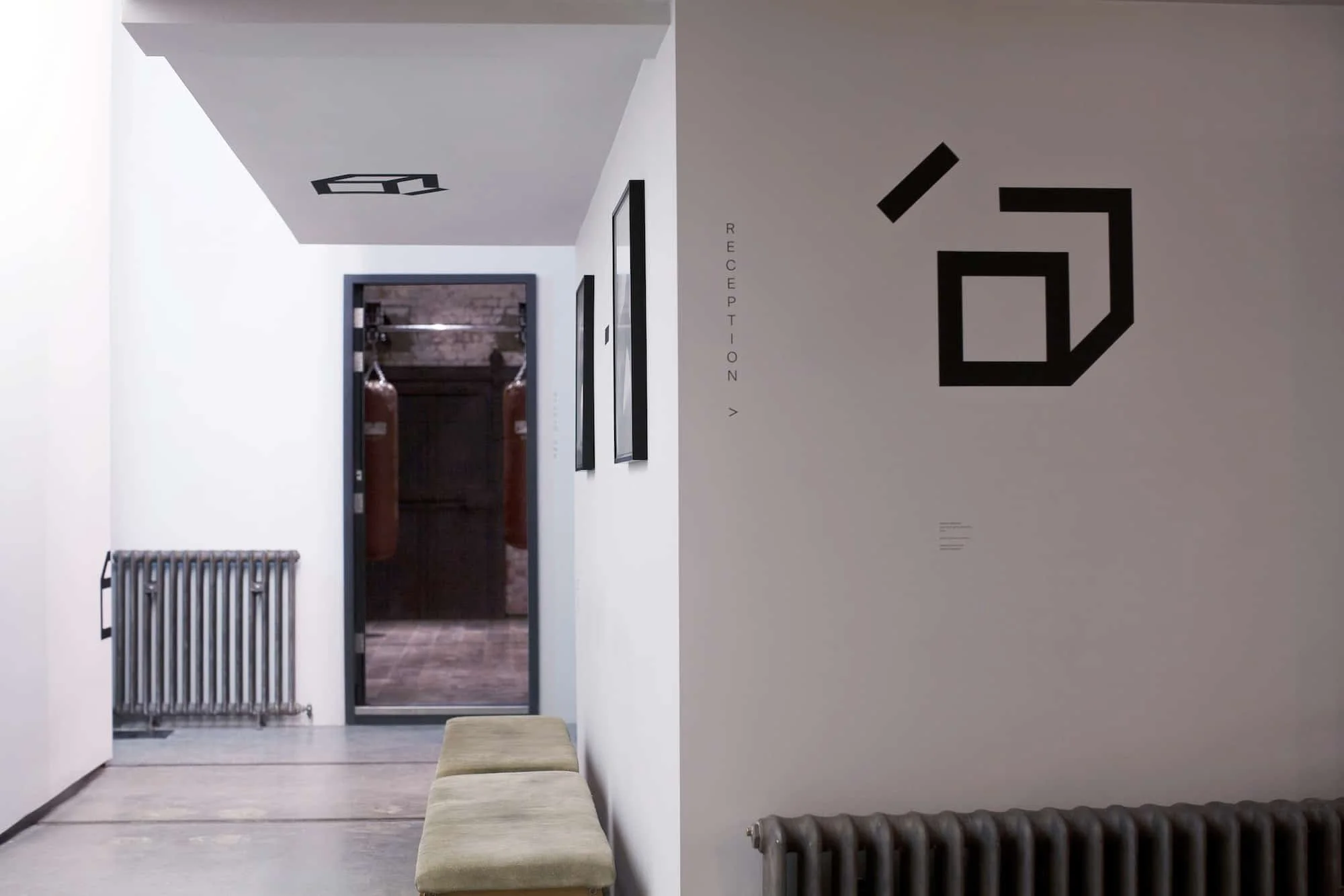 Interior of a modern building with a bench in the foreground, a white wall with black geometric art and the word 'RECEPTION' written vertically, a glass door leading to a hallway with hanging boxing bags, radiator, and framed artwork on the walls.