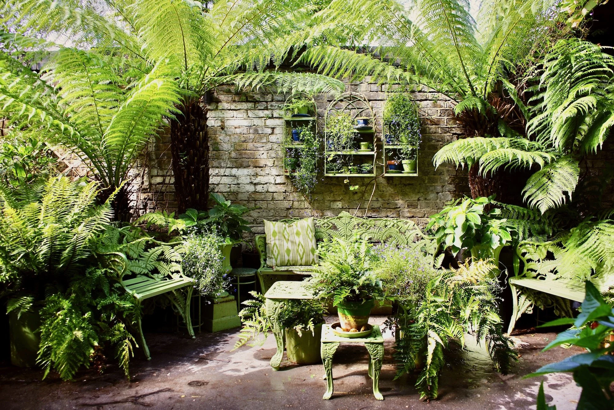 An outdoor garden seating area with vintage-style furniture surrounded by lush green ferns and potted plants, with a brick wall in the background and decorative wall shelves with plants.