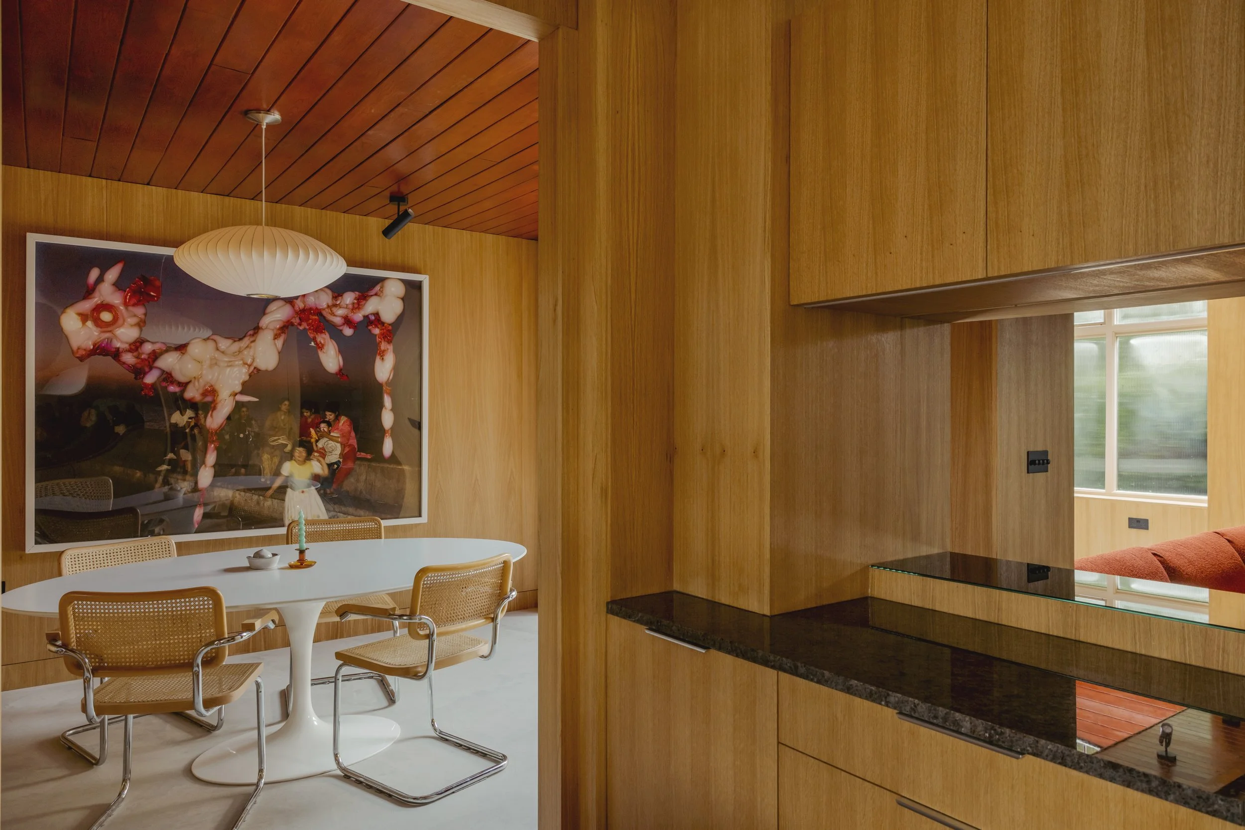 A wooden-paneled dining area with a white round table and three beige chairs. A large framed abstract artwork hangs on the wooden wall. There is a white pendant light hanging from the ceiling.