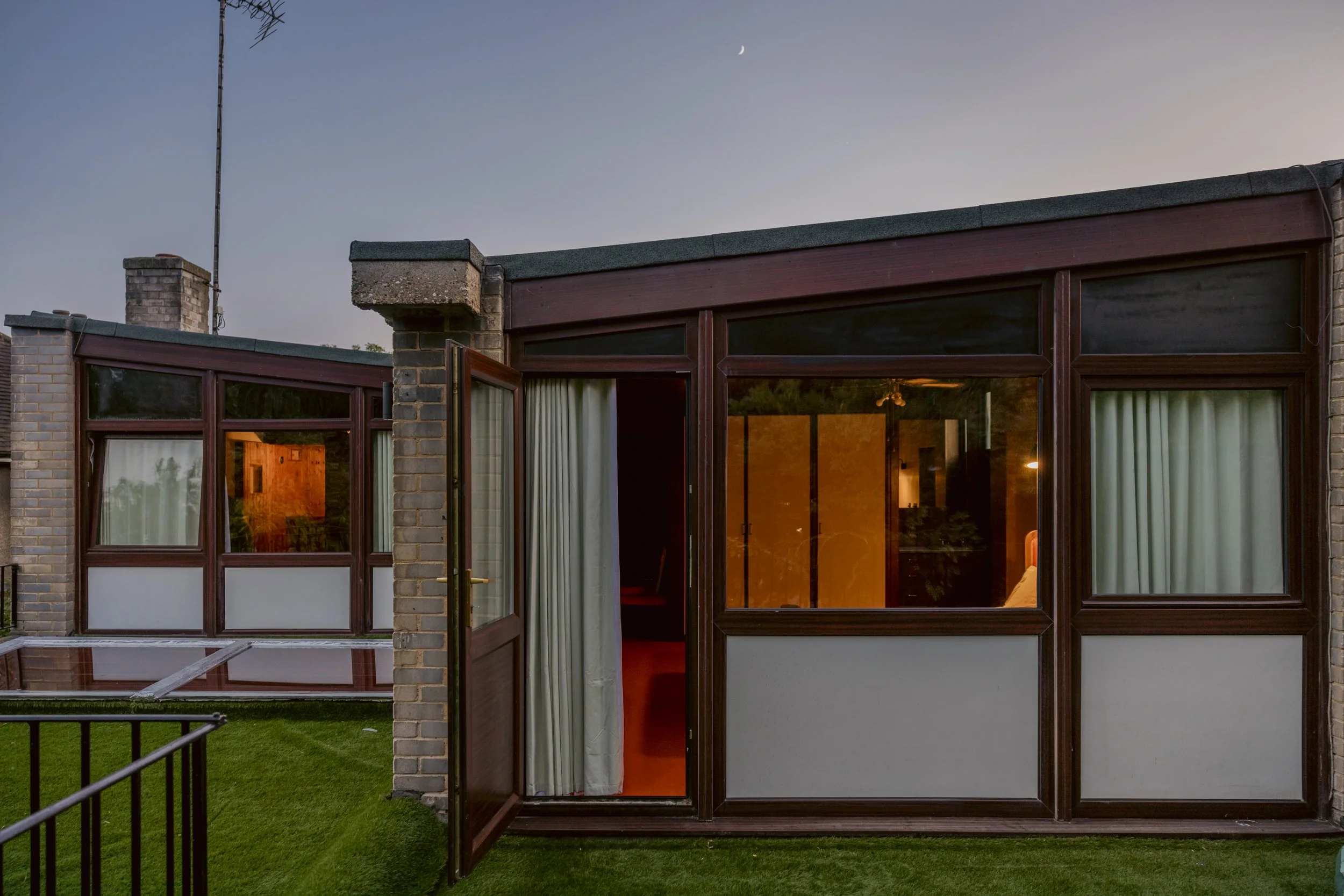 Exterior view of a residential building with large glass windows, open door, and a view of the interior at dusk. The building has a brick and wood exterior, with a lawn in the foreground and a clear sky with a visible crescent moon.