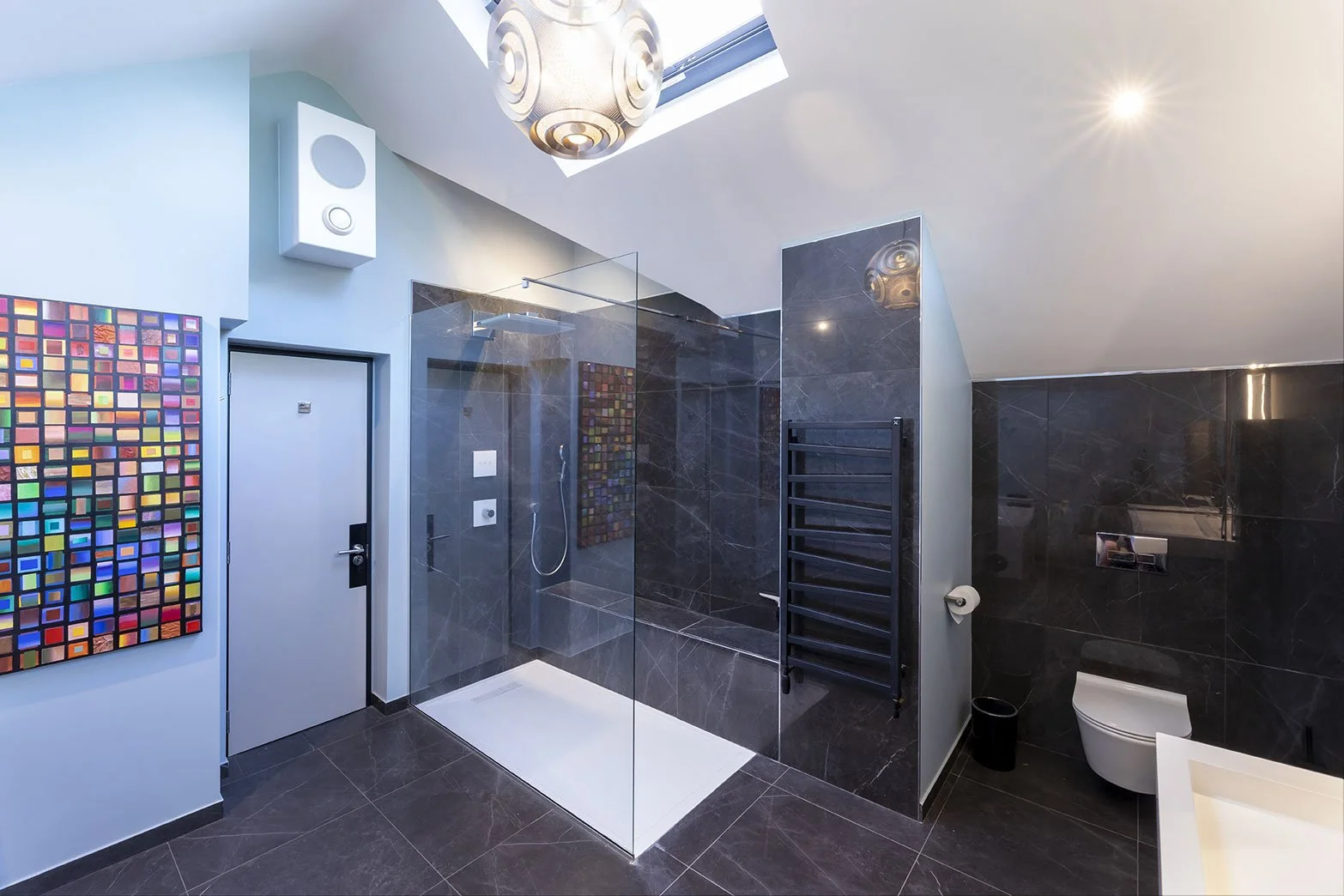 Modern bathroom with black marble walls and floors, a glass-enclosed shower, a wall-mounted toilet, a black heated towel rack, a colorful abstract art piece on the wall, a ceiling skylight, and a decorative ceiling light.
