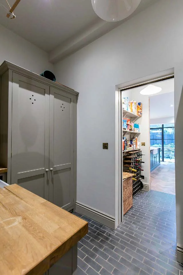 View of a door leading to a pantry with shelves stocked with food and spices, next to a woven basket and a wine rack. Part of a kitchen with a wooden countertop and gray tiled floor.