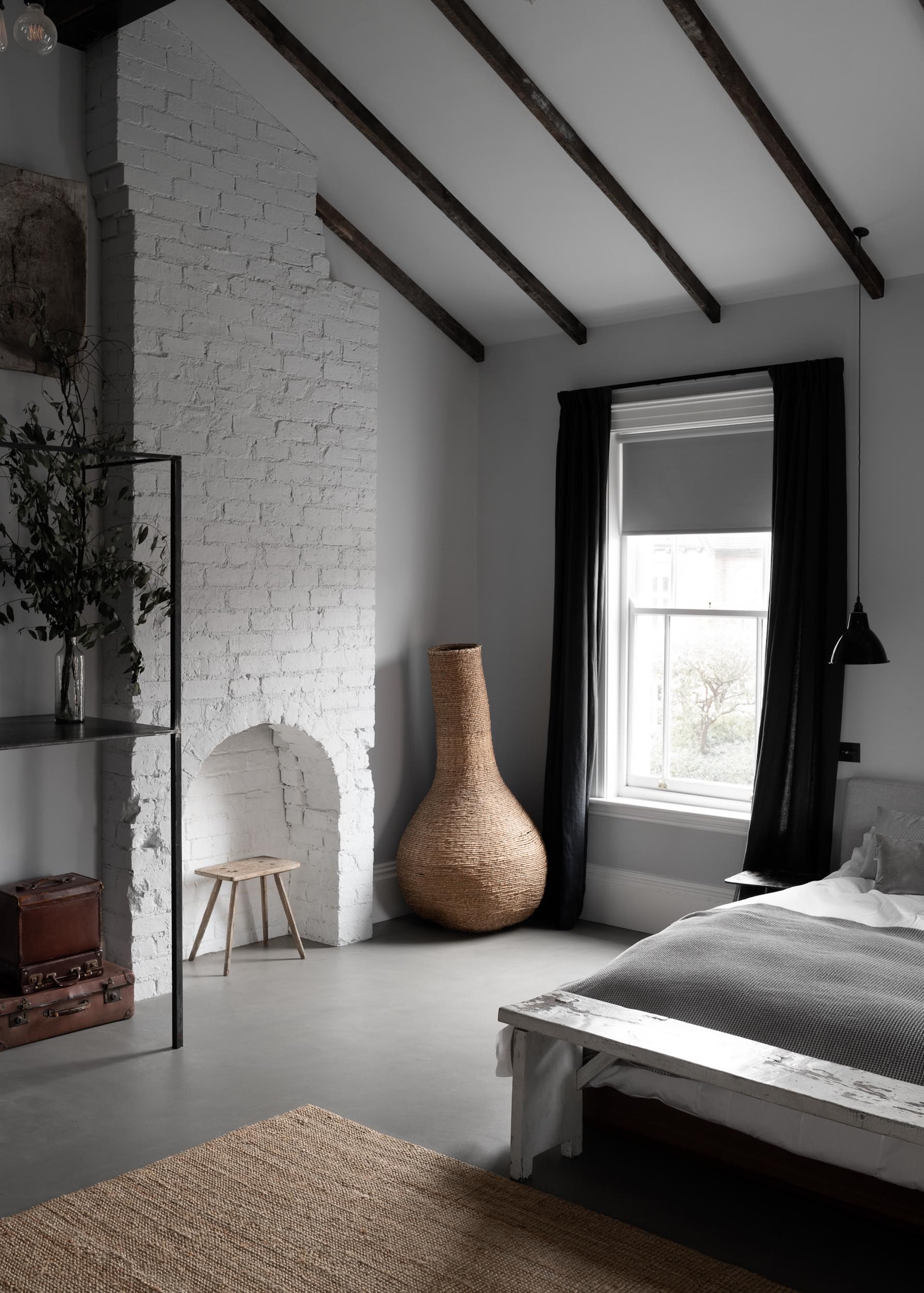 A minimalist bedroom with a white brick fireplace, large window with black curtains, and wooden beams on the ceiling. There is a short wooden stool, a large woven vase, and a bed with white and gray bedding.