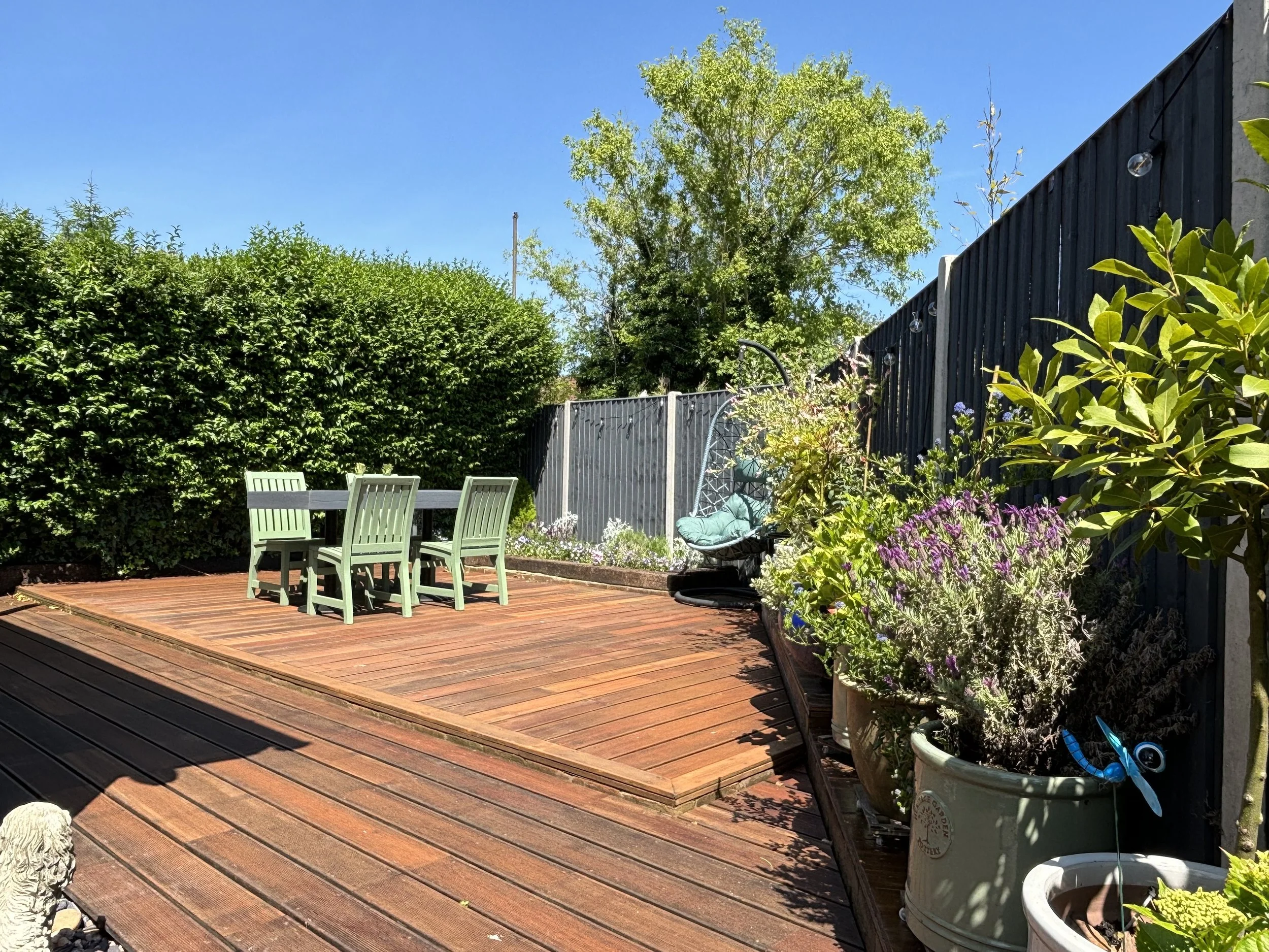 A backyard patio with wooden decking, green chairs around a table, potted plants with purple and green foliage, a black fence, and clear blue sky.