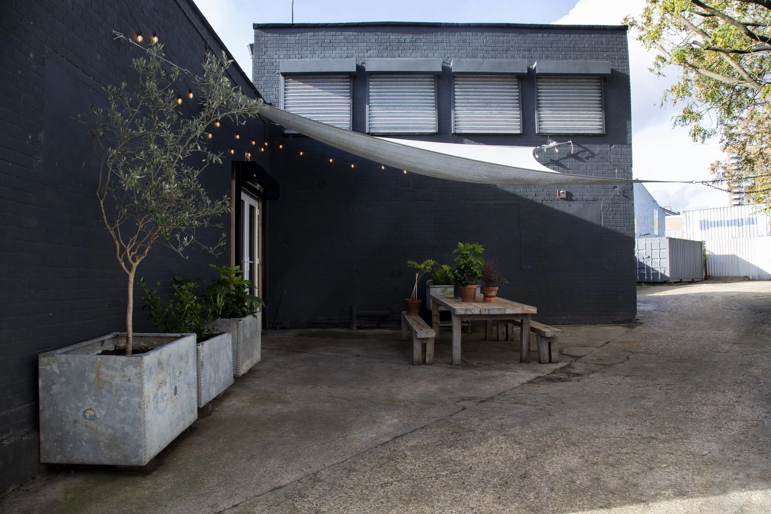 An outdoor patio area with a black brick wall, a wooden picnic table with benches, potted plants on the table, and a string of lights overhead. There is a large grey shade sail above, and the ground is concrete. A small tree in a concrete planter is 
