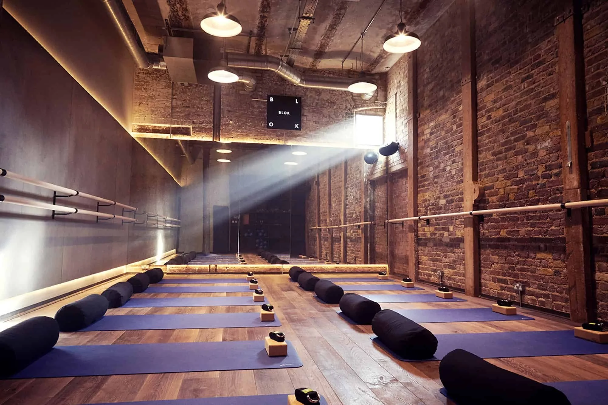 An empty yoga or fitness studio with purple mats, black bolsters, and wooden blocks arranged on a wooden floor. Sunlight streams in through a window, creating a beams of light. The studio has exposed brick walls and industrial ceiling lighting.