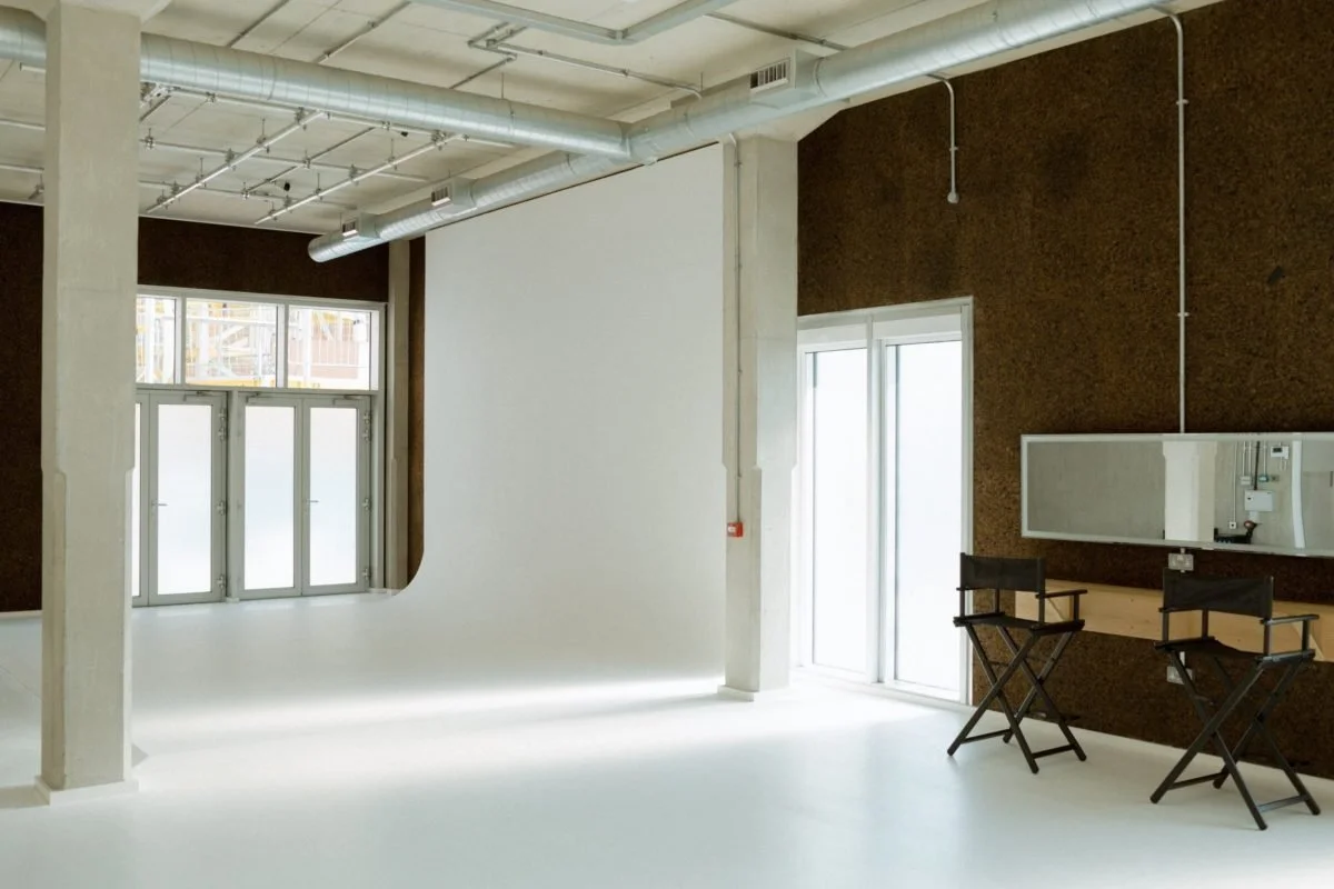 Empty modern interior space with large windows and doors, dark brown textured wall, white floor, and two director chairs near a small counter with a mirror.