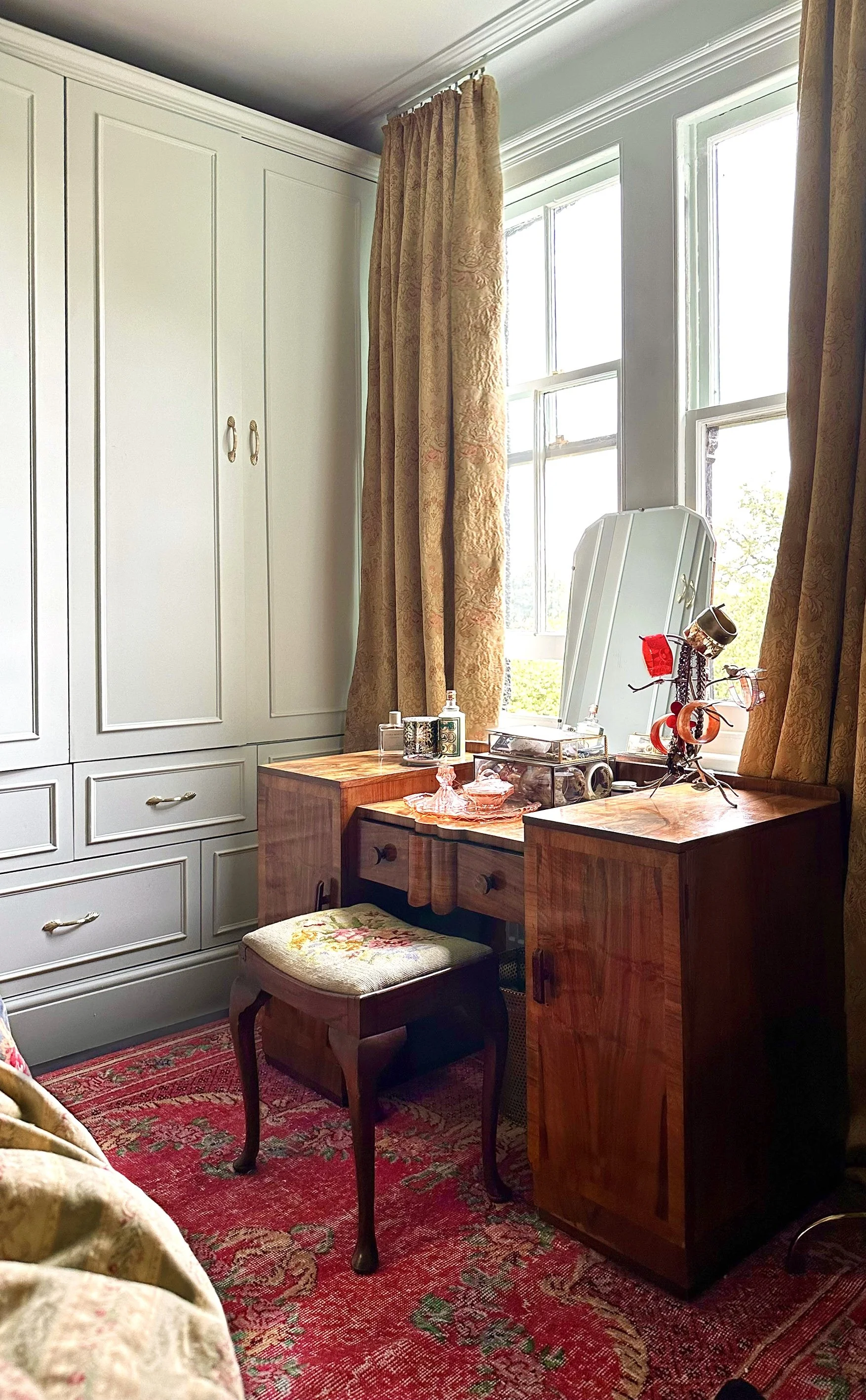 A vintage-style room corner with a white wardrobe, a wooden vanity table, a cushioned stool with floral embroidery, and a red patterned rug. The room has two windows with gold curtains, and the vanity table holds assorted glassware, bottles, and deco