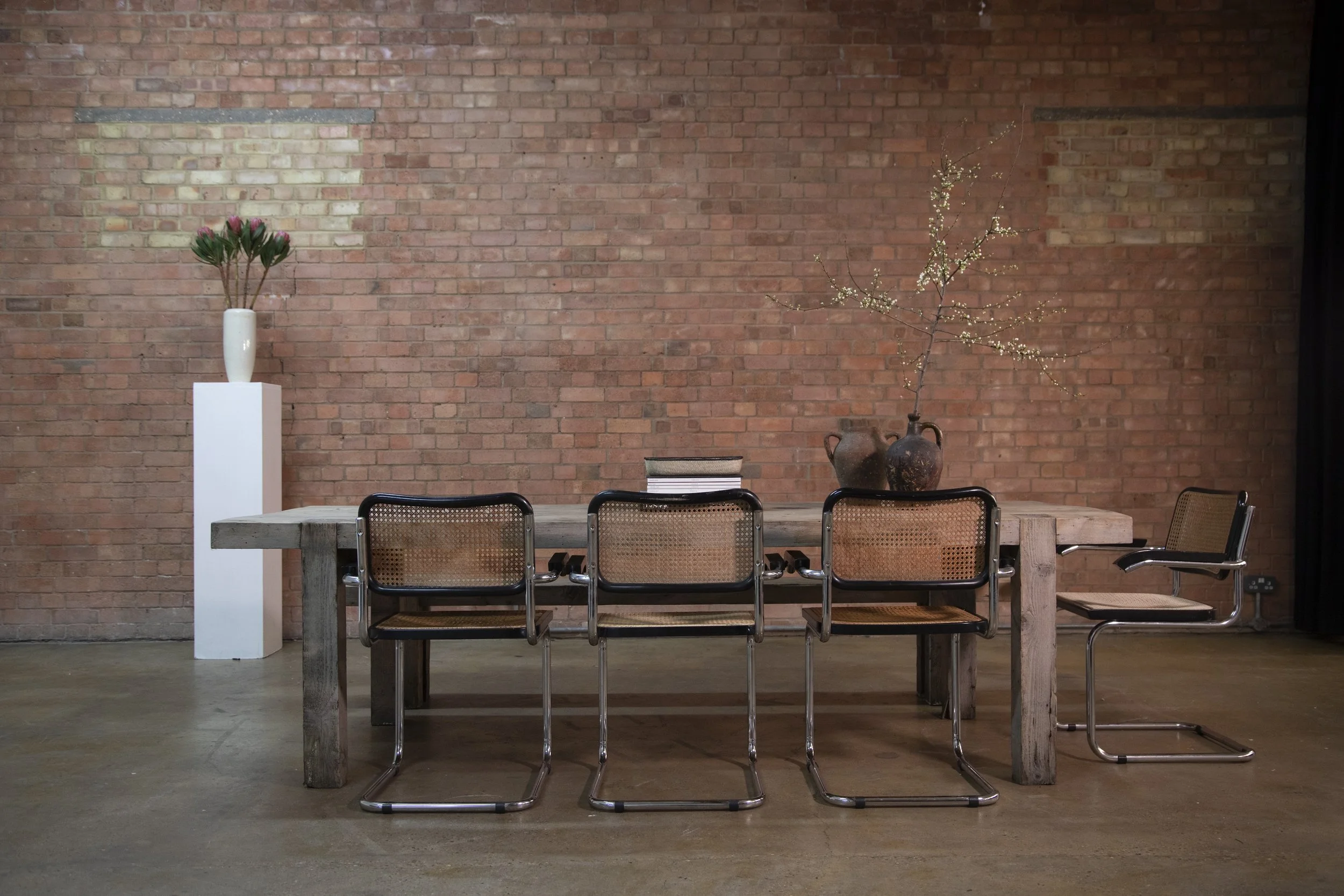 A rustic wooden dining table with four modern chairs against a brick wall, decorated with two flower arrangements in vases and a stack of books.