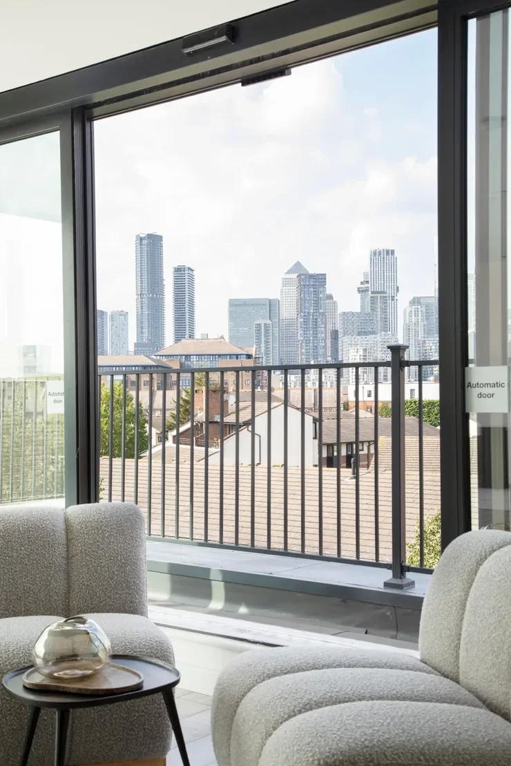 Living room with large open glass doors, view of city skyline with tall buildings, small balcony with black railing, and modern chairs with a decorative item on a small table.