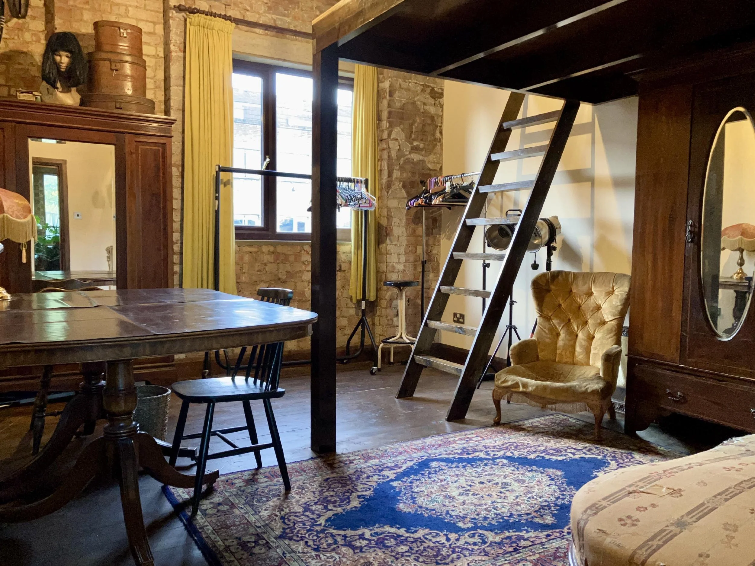 A cozy living space with vintage furniture, a wooden table, a black chair, a mustard-colored tufted armchair, a wooden wardrobe with an oval mirror, a staircase with a ladder leading to a loft, a window with yellow curtains, and various collectibles 