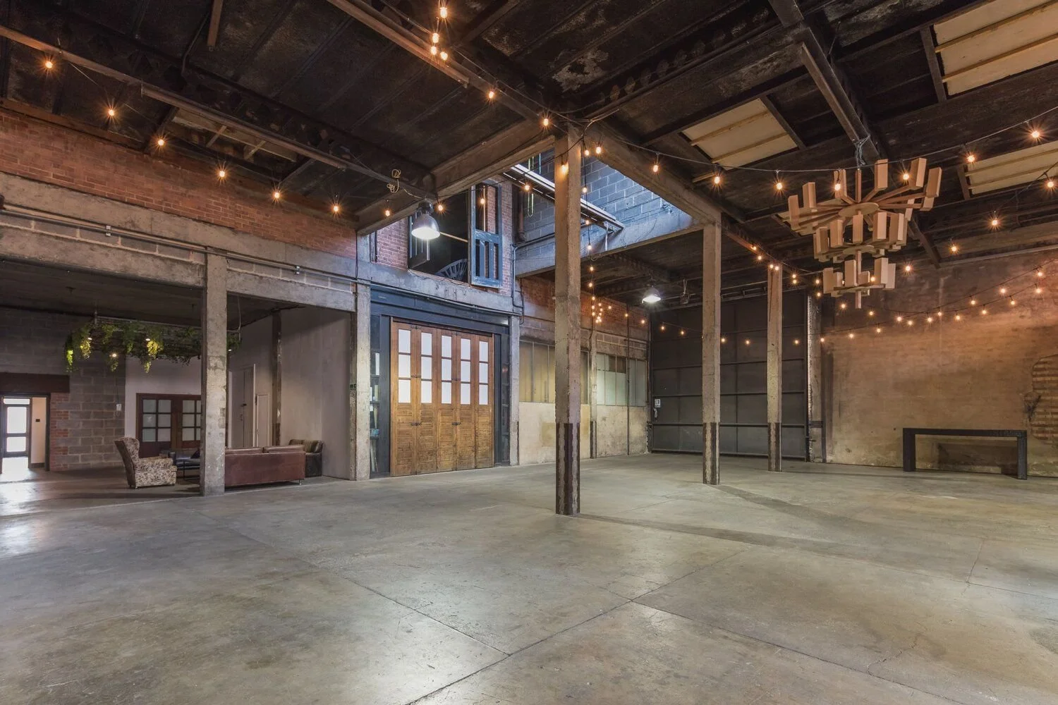 An empty industrial-style event space with exposed brick and concrete walls, concrete flooring, and string lights hanging from the ceiling. There are some seating areas with couches in one corner and a large wooden sliding door in the center.