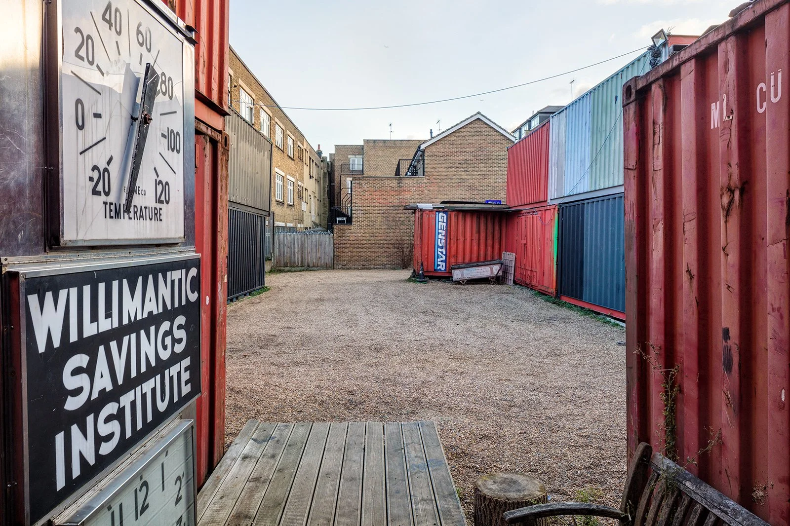 An outdoor area with colorful shipping containers forming a fence, a weathered wooden bench, and a gravel ground. In the background, there are brick buildings and a clear sky.