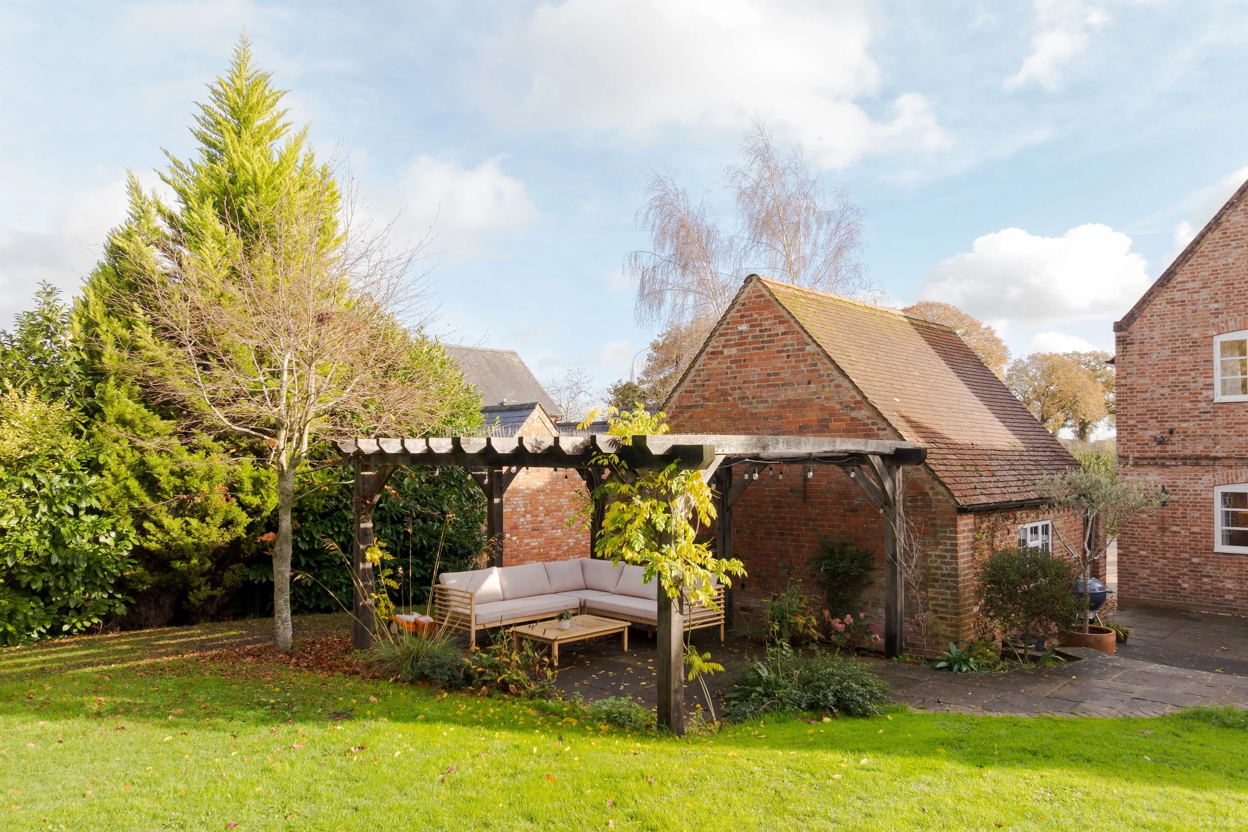 Backyard garden with grassy lawn, a small tree, and a wooden pergola with a white outdoor sectional sofa beneath it, surrounded by various plants and bushes, with brick houses and a blue sky with clouds in the background.