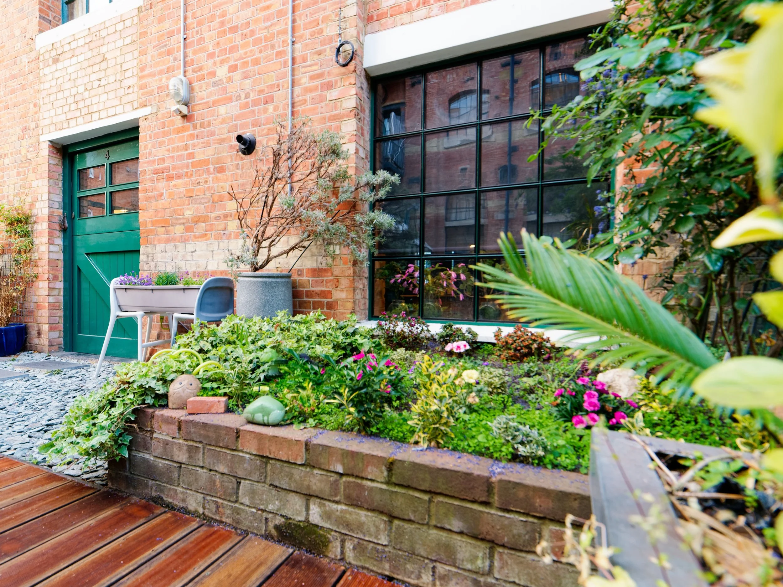 A backyard garden with a brick wall, large window, potted plants, and colorful flowers, with a wooden deck in the foreground.