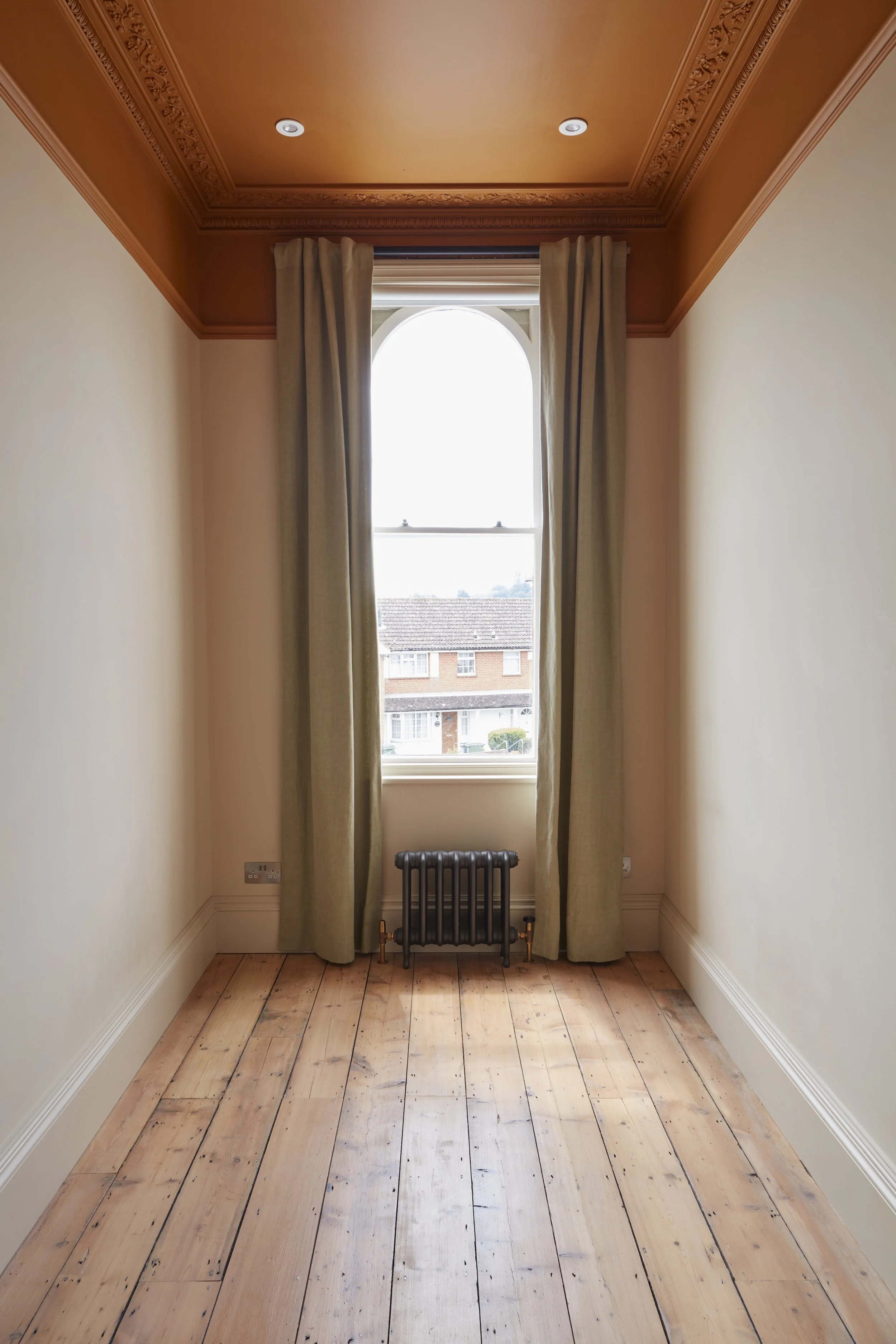 An empty room with light-colored walls, a wooden floor, a large arched window with beige curtains, decorative crown molding, and small recessed ceiling lights.