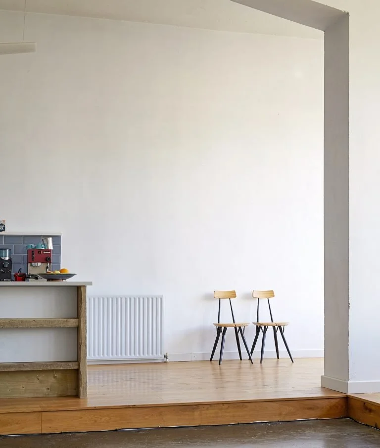 Part of a kitchen with a countertop on the left, a white radiator on the wall, and two wooden and black chairs against a plain white wall in the background.