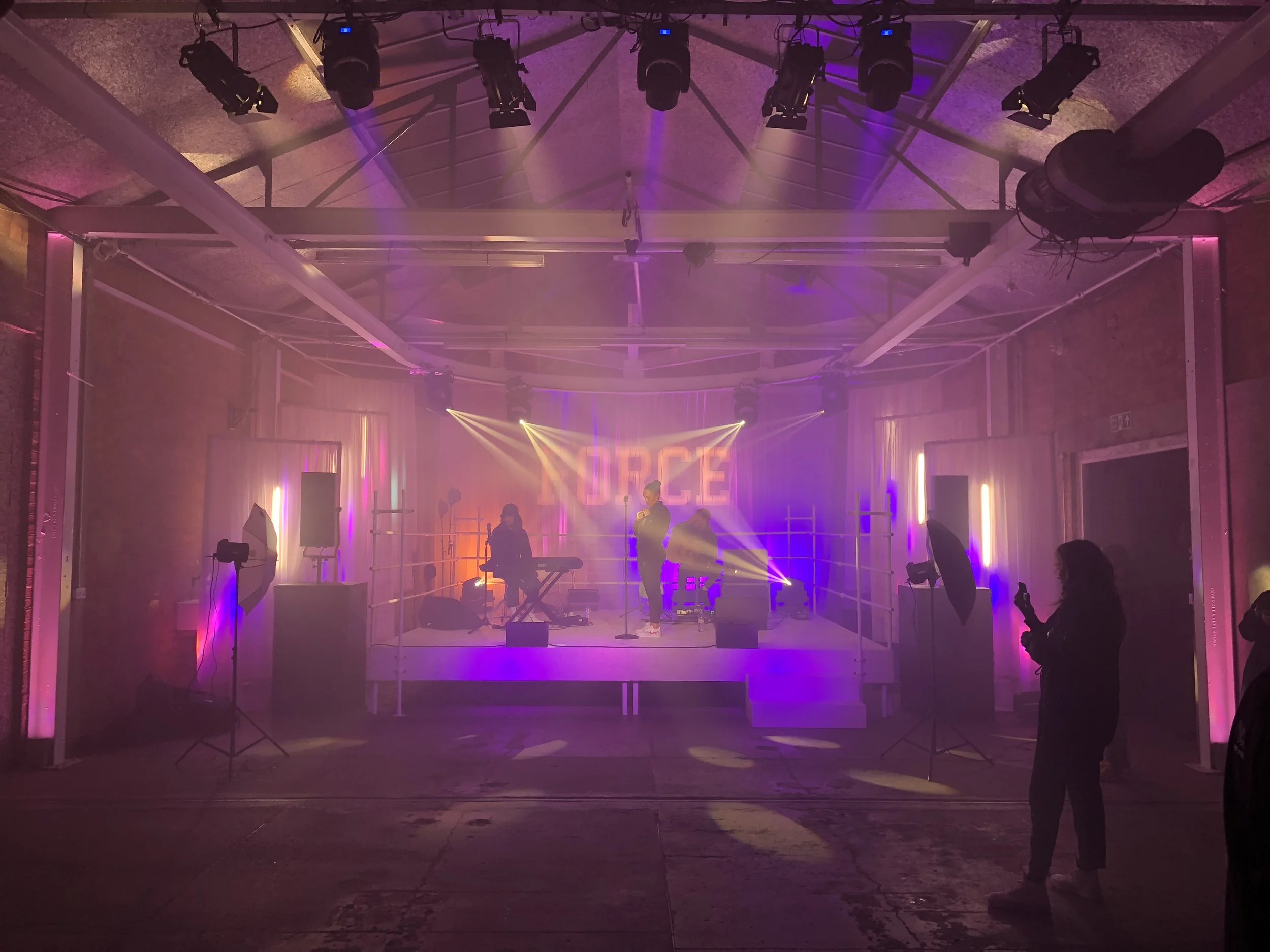 Stage with three standing performers and a keyboard player, colorful lighting, pink and purple hues, the word 'NOPE' projected on the background, audience members standing nearby, some holding phones, in an indoor venue with exposed brick walls and c