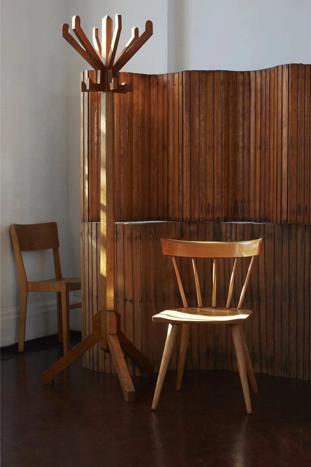A wooden coat rack, two wooden chairs, and a curved wooden privacy screen in a room with dark hardwood flooring and a light-colored wall.