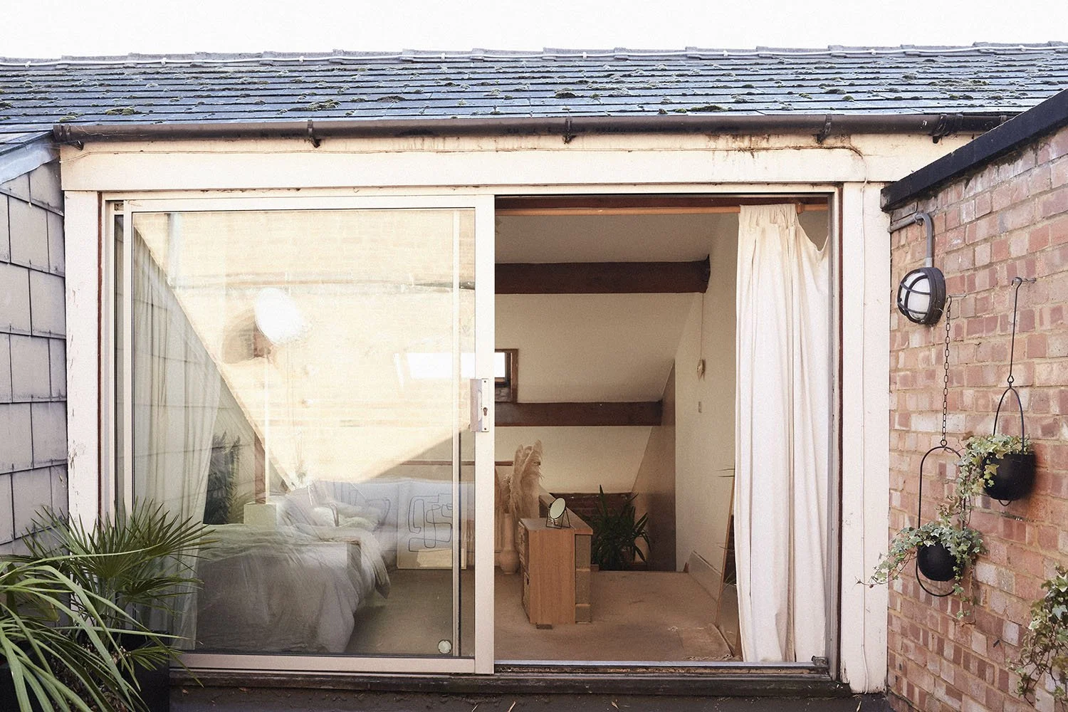 The exterior of a house showing large glass sliding doors partly covered with white curtains, revealing a living room with a grey sofa, some plants, and a bookshelf inside.