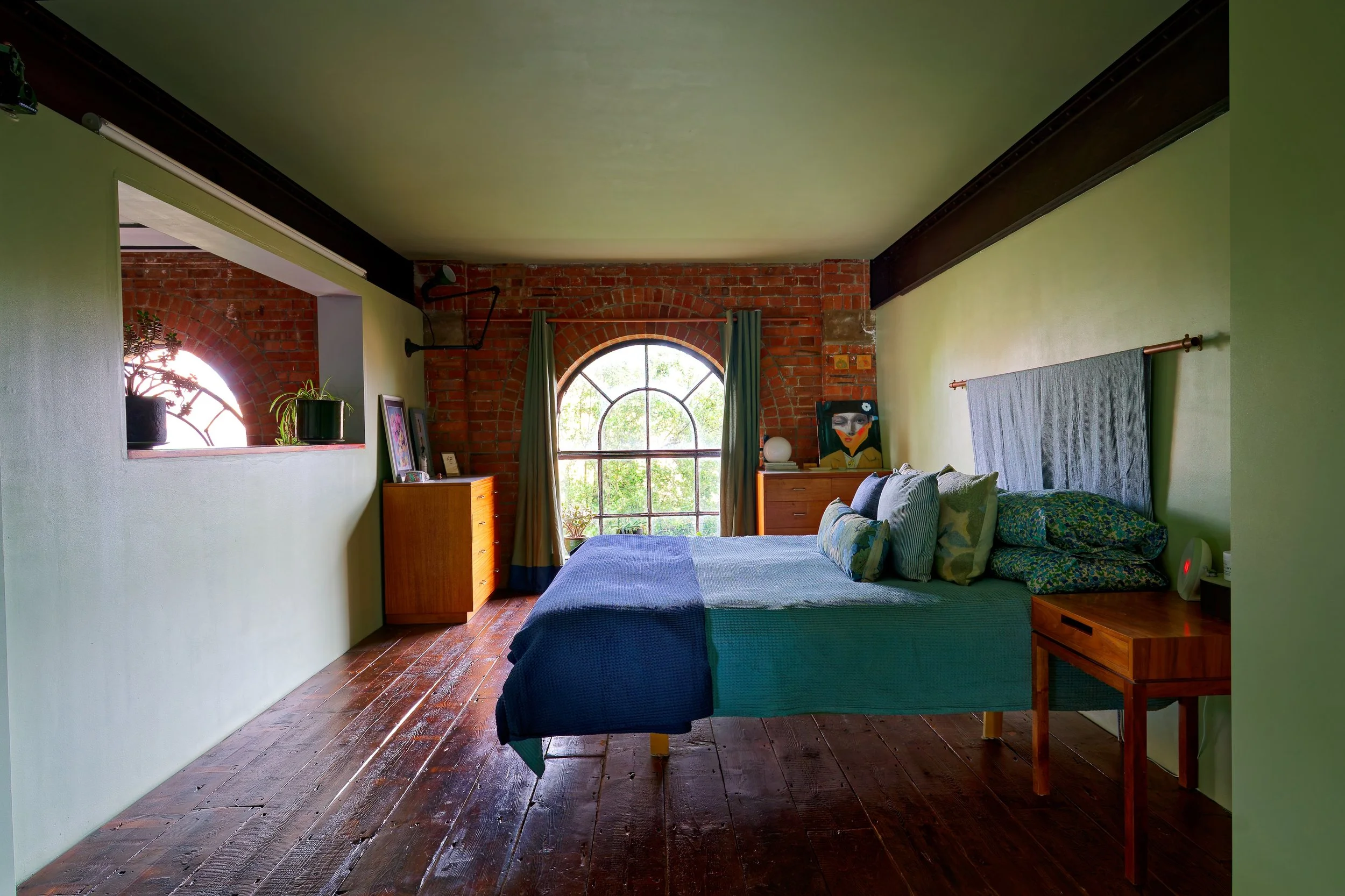 A bedroom with an exposed brick wall and large arched window with green curtains, wooden floor, bed with blue and green bedding, nightstand, and art pieces.