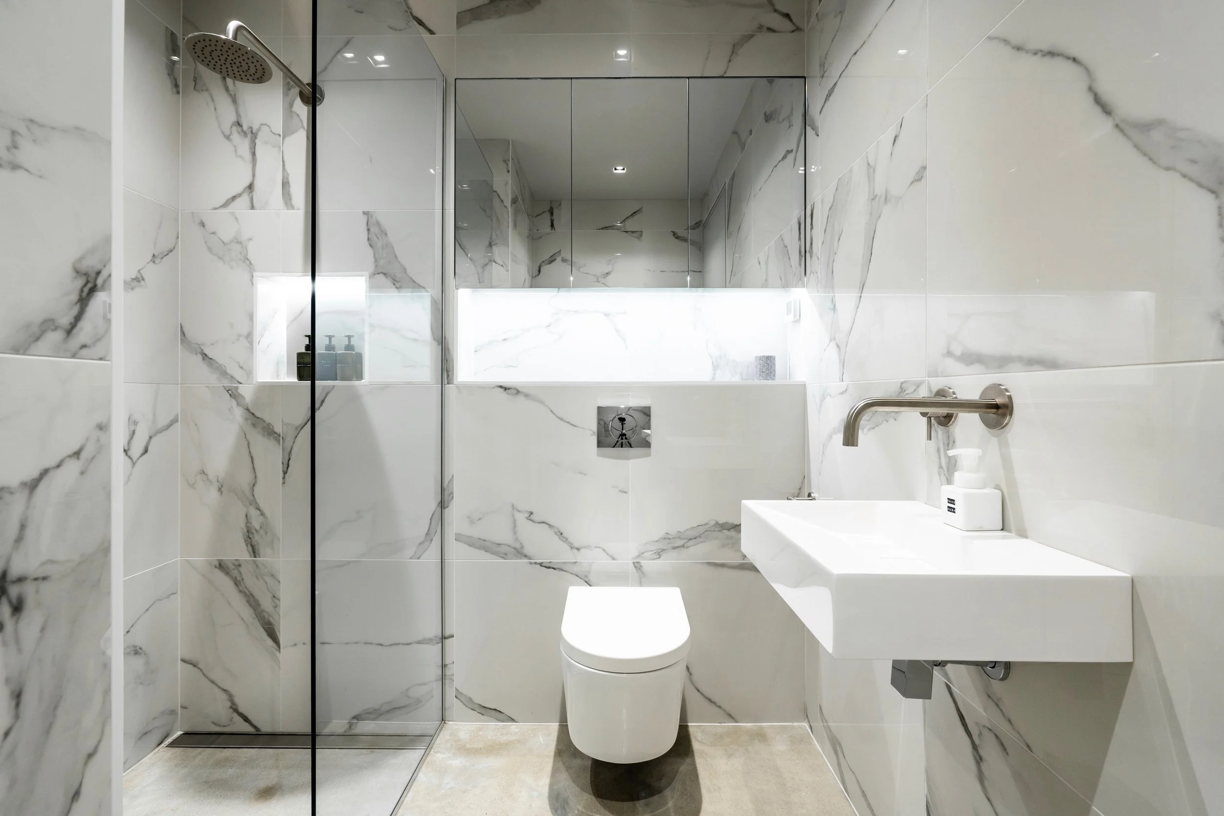 Modern bathroom with marble walls, a walk-in shower, a wall-mounted toilet, and a floating sink with a soap dispenser.