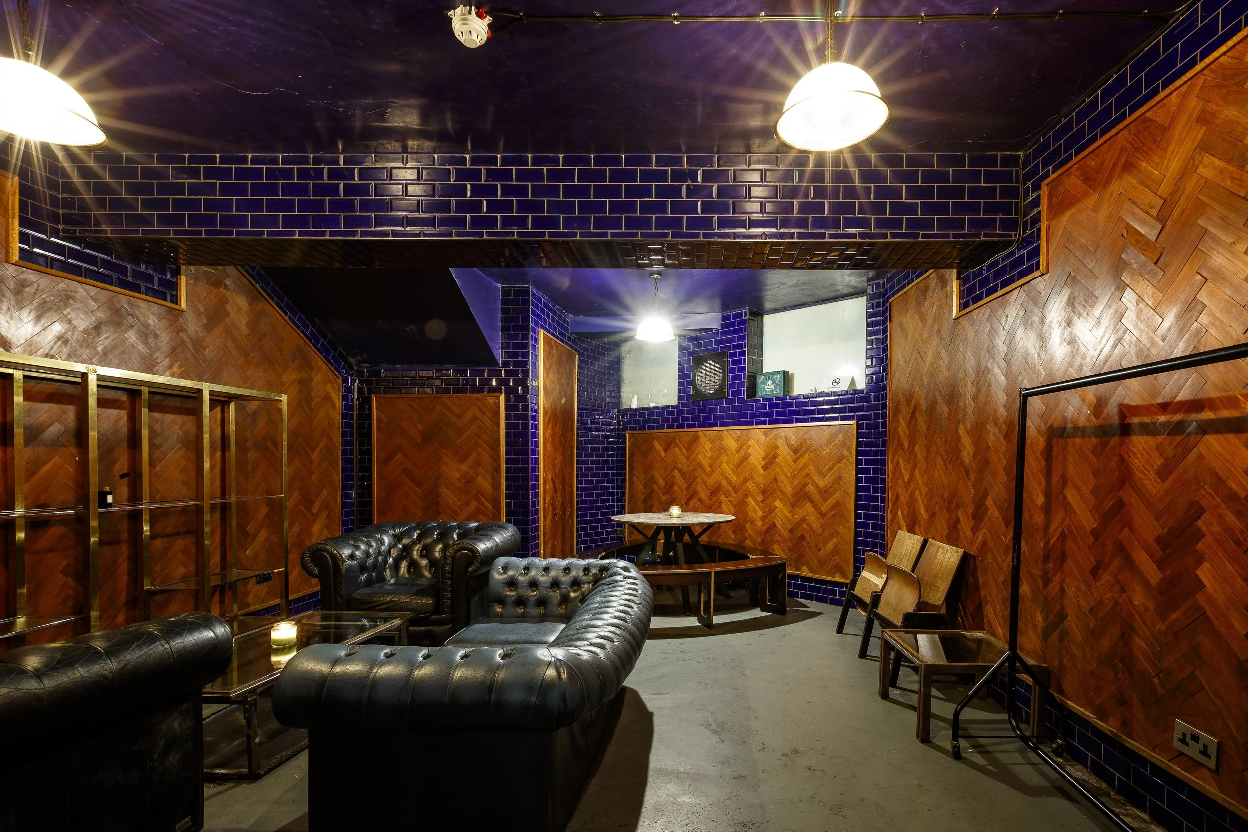 Interior of a dimly lit lounge with black leather sofas, wooden chairs, and purple tiled walls.