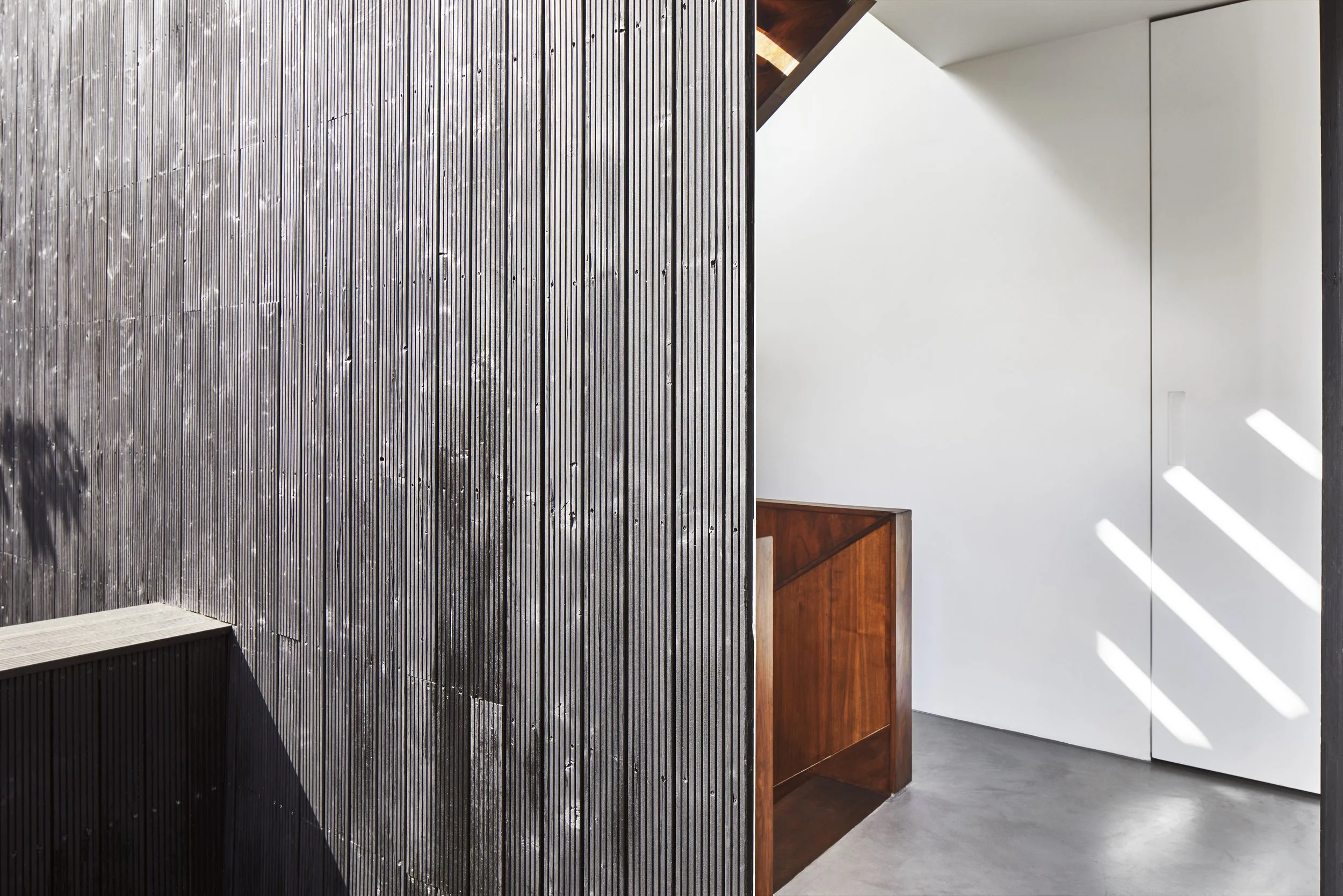 Interior view of a modern minimalist room with a textured, dark vertical wooden slat wall, a white wall with sunlight patterns, and a small wooden desk or shelf with a dark finish.