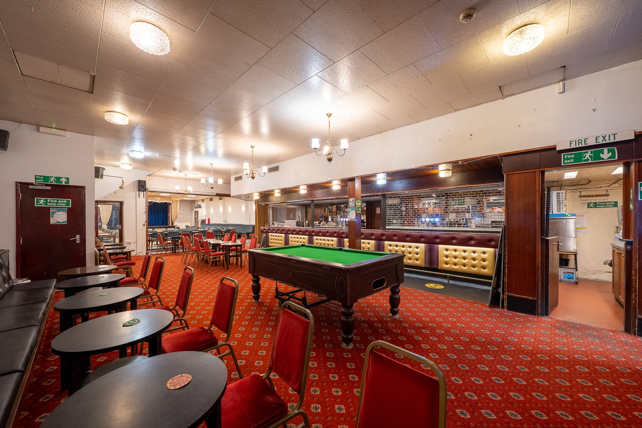 Interior of a recreational room featuring a pool table, red chairs, and tables in a casual setting, with fire exit signs and ceiling lights.