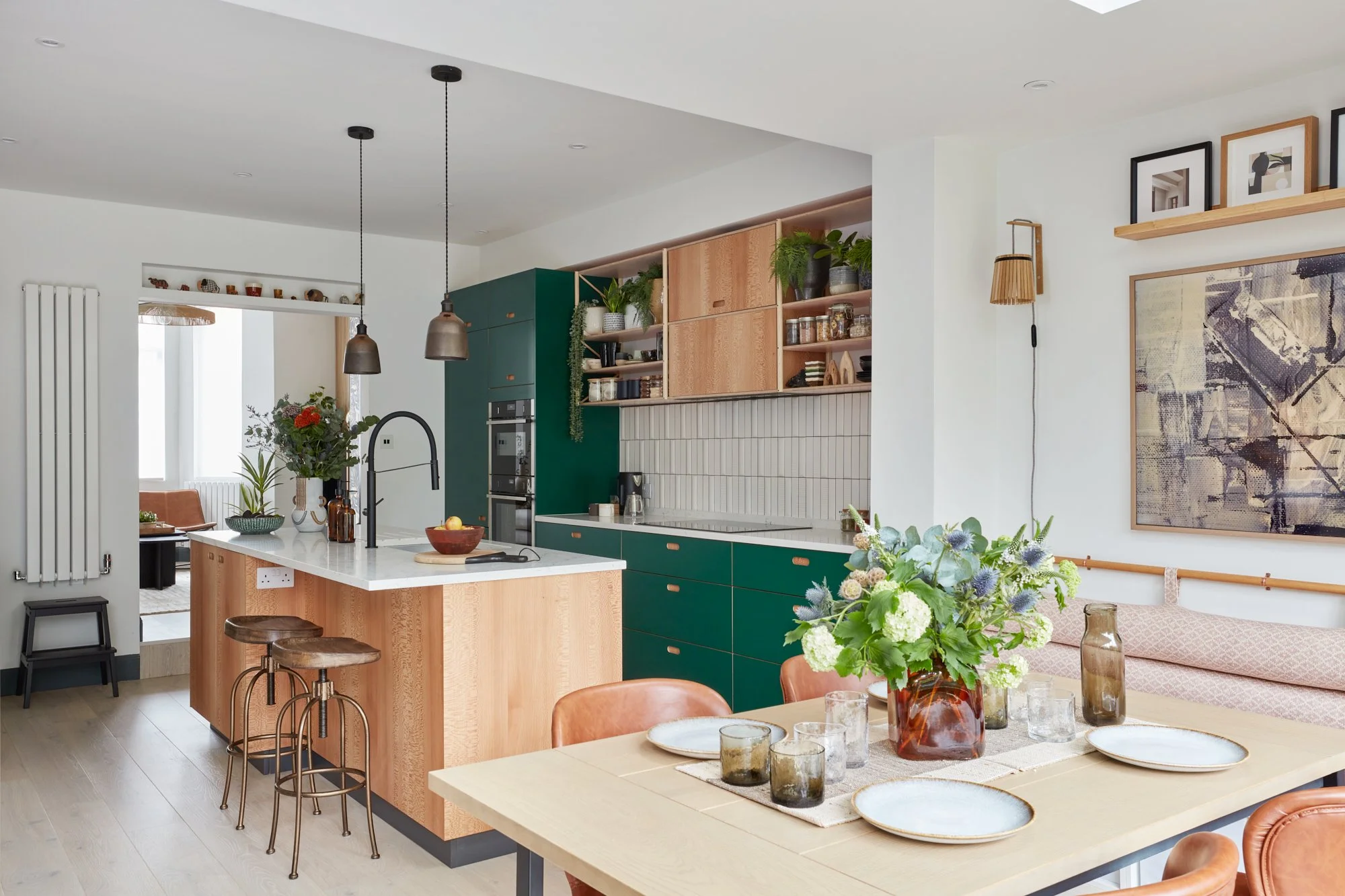 Open-concept kitchen and dining area with green and wooden cabinetry, a white island with a black sink, hanging pendant lights, and a wood dining table with a large floral centerpiece and tableware.