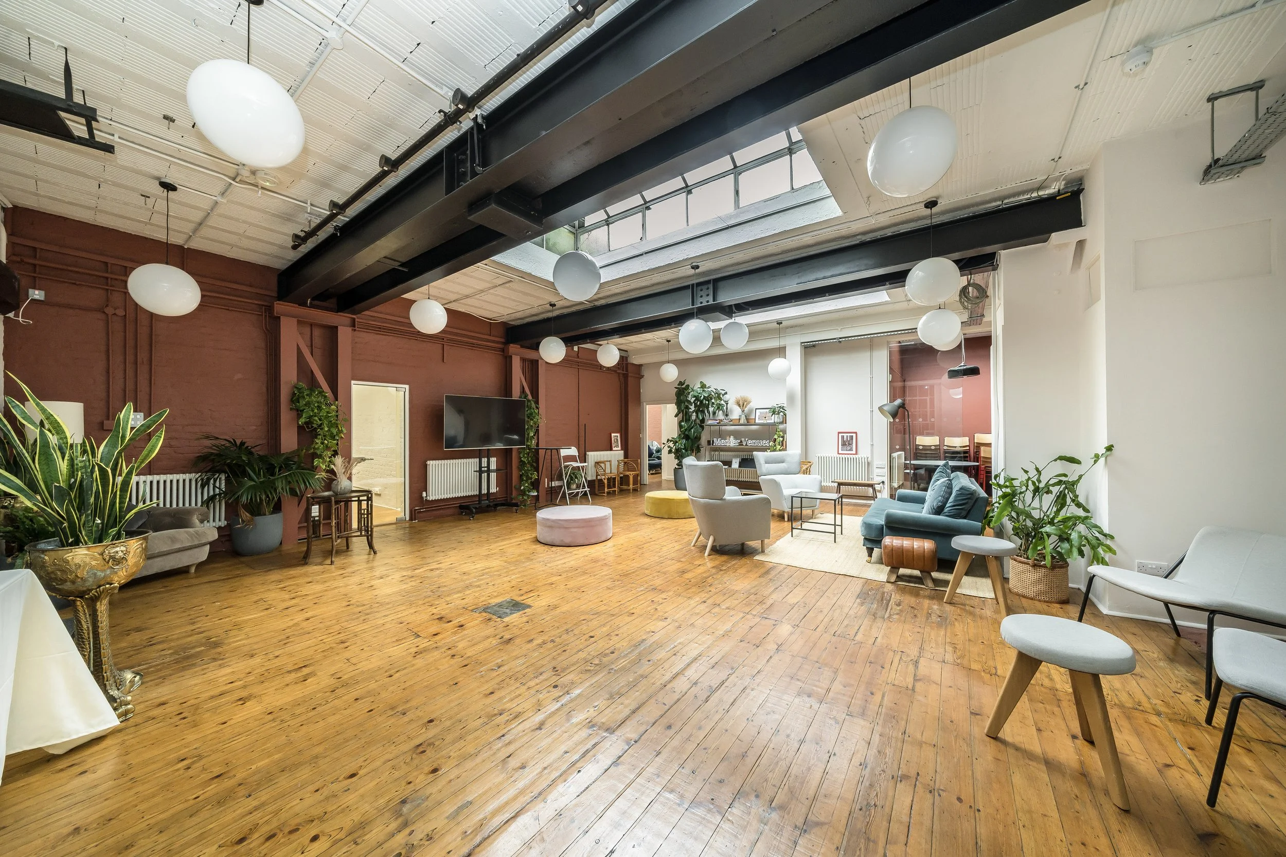A spacious lounge area with wooden floors, white and pastel-colored chairs, a large flat-screen TV, and various potted plants. The ceiling has exposed beams and round white pendant lights, with a skylight allowing natural light.