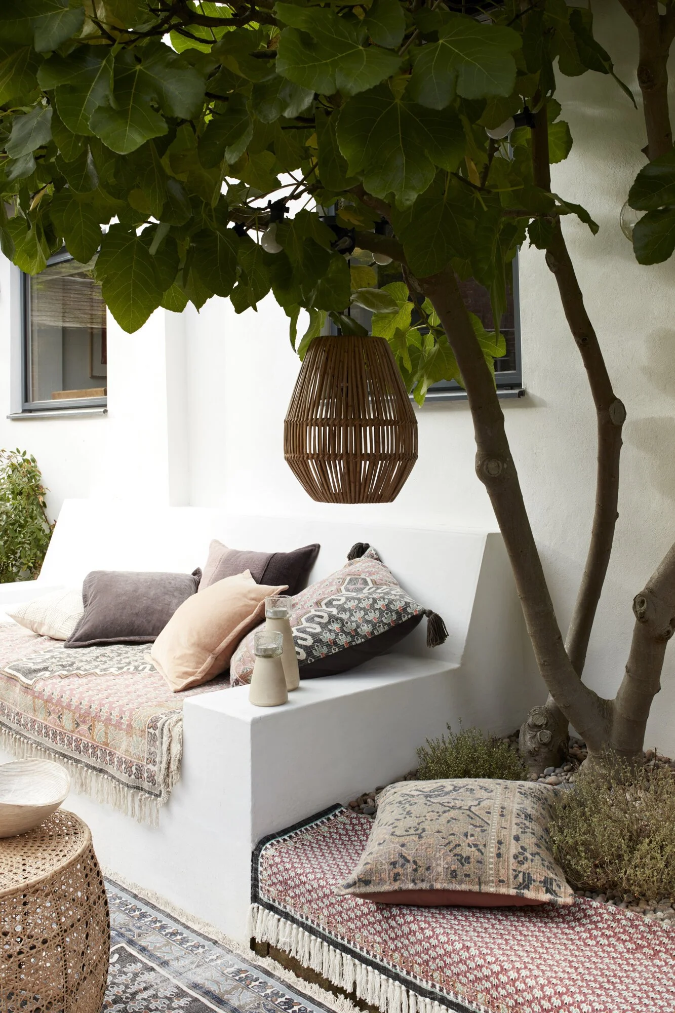 Outdoor patio area with a white built-in bench decorated with patterned pillows, a woven area rug, small vases, a wooden hanging lantern, a tree with large green leaves, and a window on a white wall.