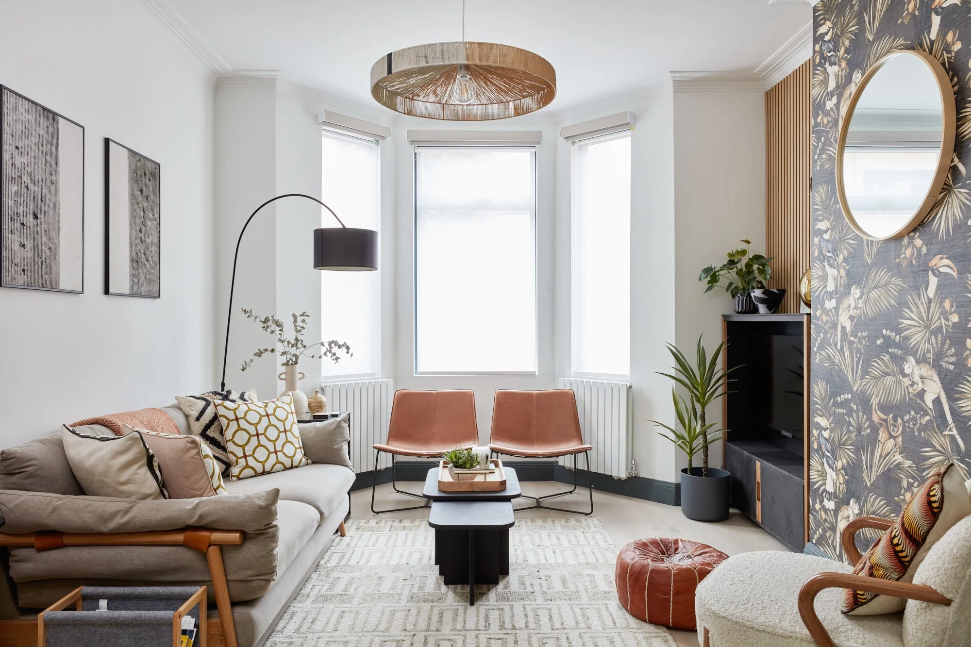 A modern living room with a beige sofa, peach chairs, a black TV stand, candlestick, and built-in window seating. There's a round ceiling light, black arc lamp, and decorative wallpaper with a tropical theme on one wall. Potted plants and framed art 