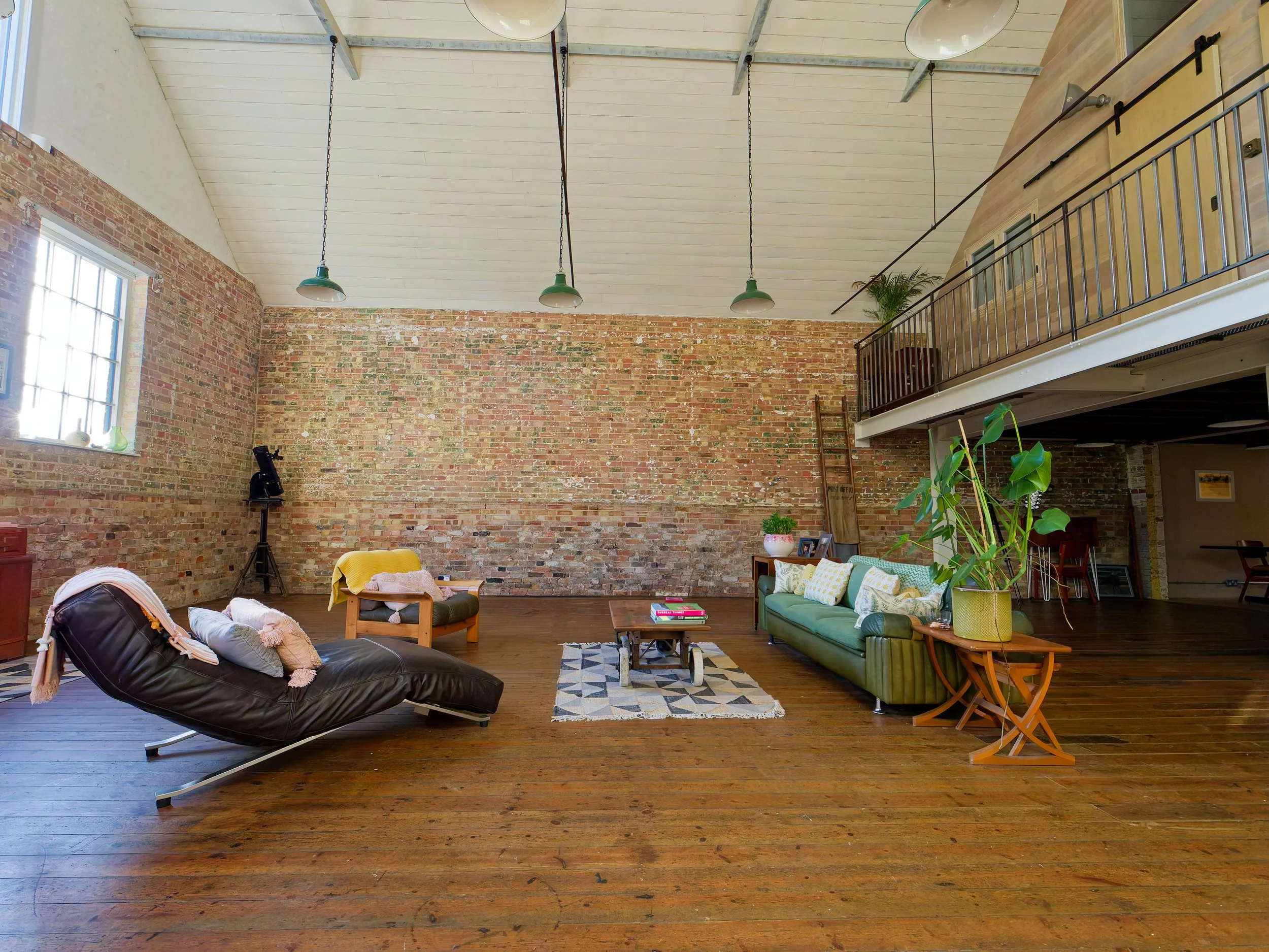 Open living room with brick wall, wooden floors, modern furniture, large windows, potted plants, and a loft area