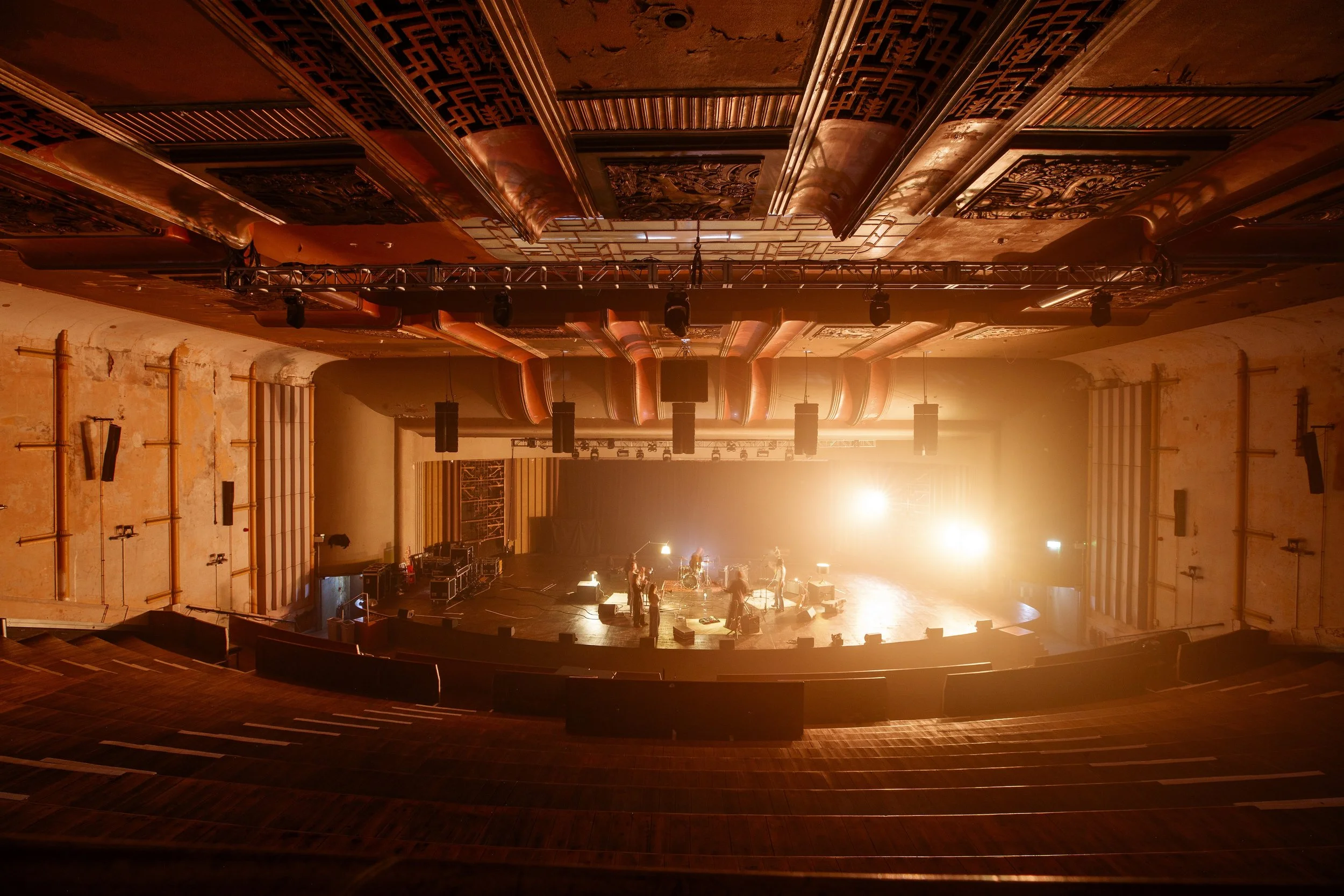 An empty theater stage with musical instruments and equipment illuminated by bright stage lights, with the audience seats in the foreground.