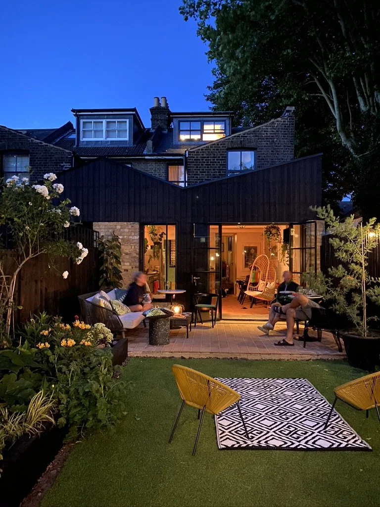 Backyard patio at dusk with three people sitting on outdoor furniture, plants, and a black-and-white rug with yellow chairs in the foreground, and a house with warm interior lighting visible through large windows.